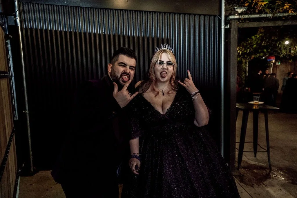Two people posing at an outdoor event, both making rock sign gesture with excited expressions, one dressed in black and the woman in a sparkly dark gown with glasses and a small tiara.
