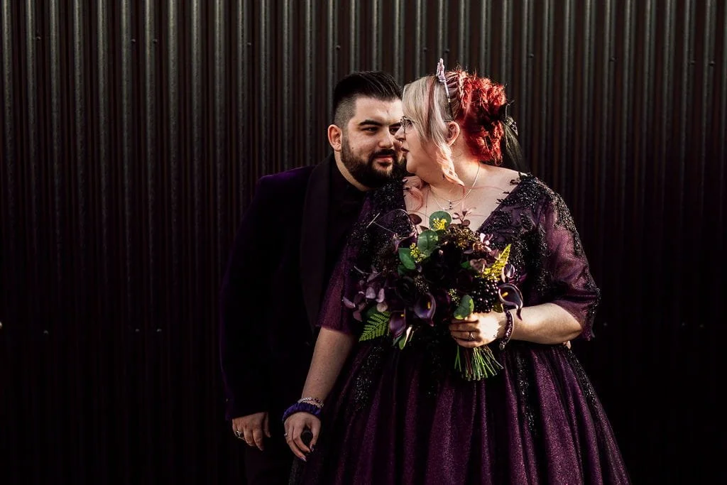 Couple dressed in dark attire, standing close with foreheads touching, in front of black corrugated metal wall. The woman holds a bouquet of dark-colored flowers and greenery.
