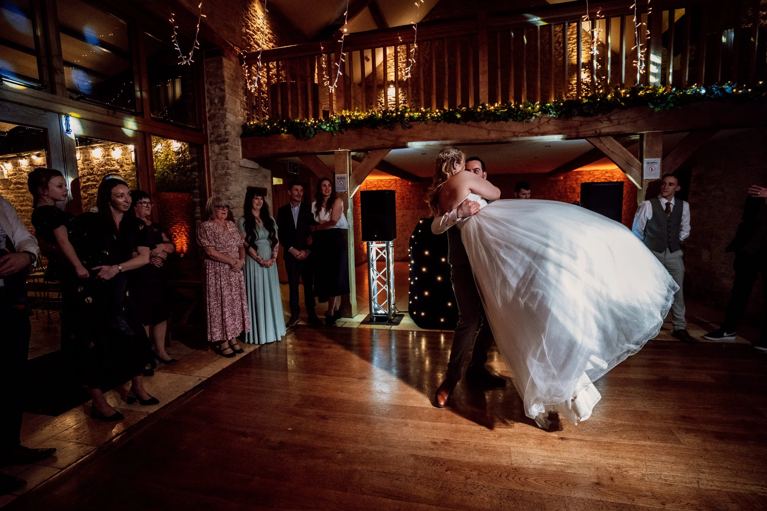 Bride in white wedding dress being dipped by groom during wedding dance, guests watching in a warmly lit rustic venue with wood and brick decor.