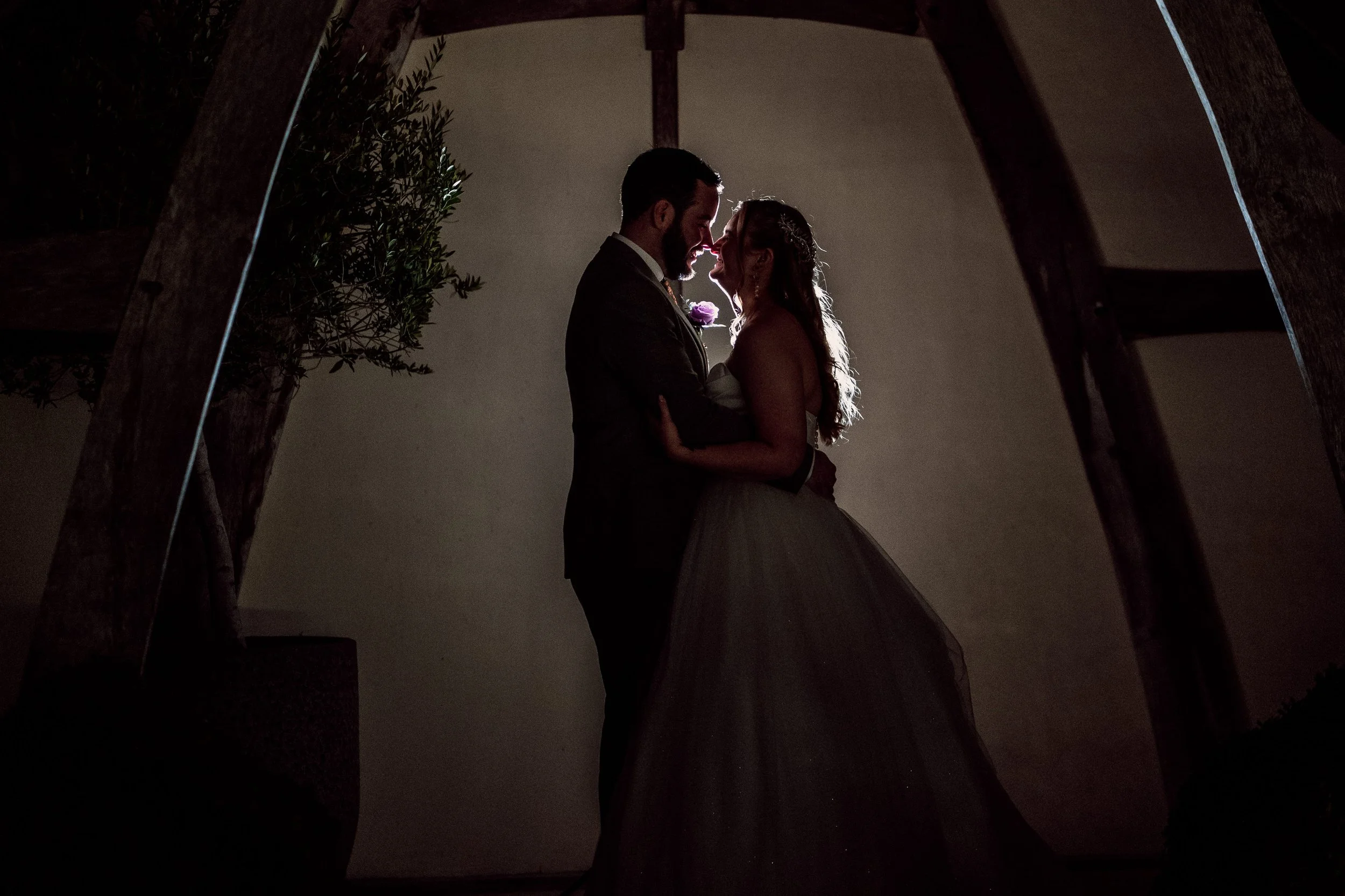 Silhouette of a bride and groom standing close together inside a wooden arch, touching foreheads in a romantic moment with backlighting.