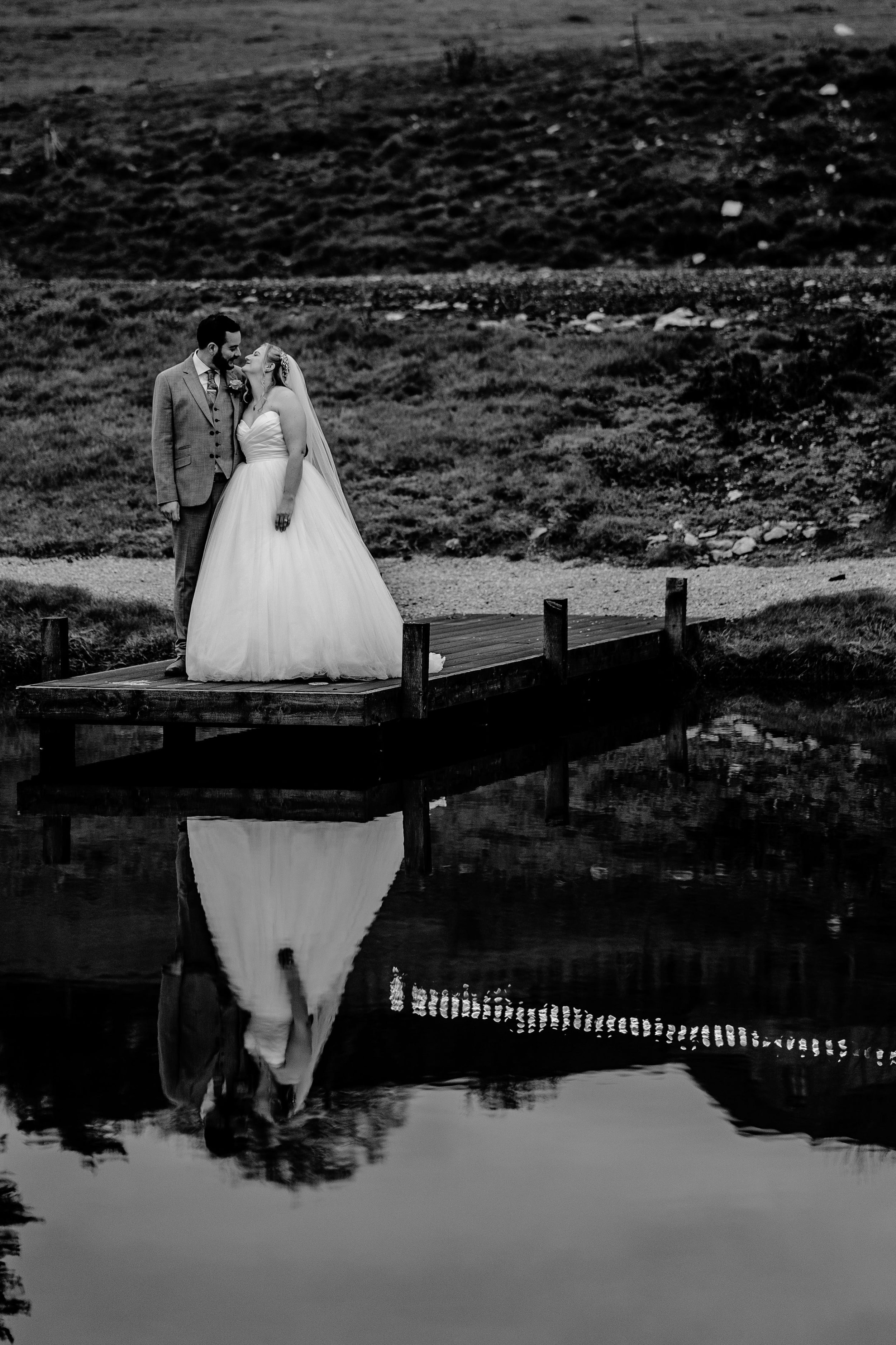 Black and white photo of a bride and groom standing on a small wooden dock by a pond, embracing and smiling at each other, with their reflection visible in the water.