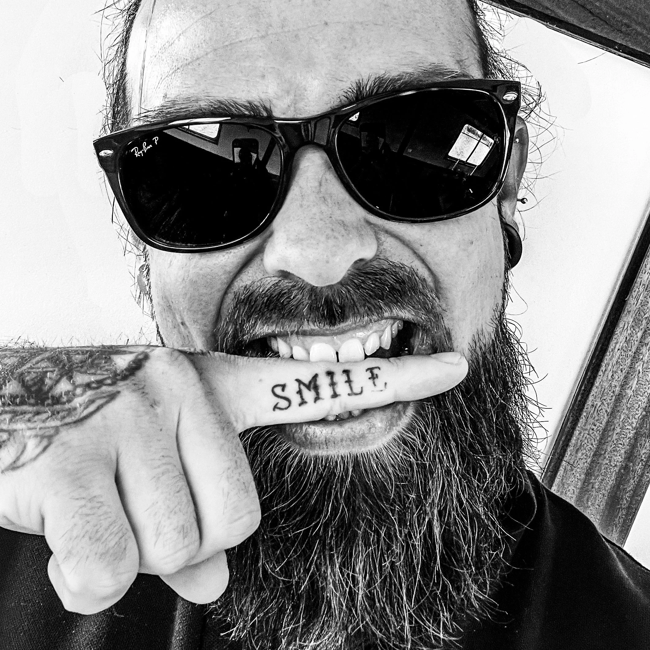 A man wearing sunglasses and smiling, showing his teeth, with a long beard and tattooed hand that has the word 'SMILE' written on his finger.