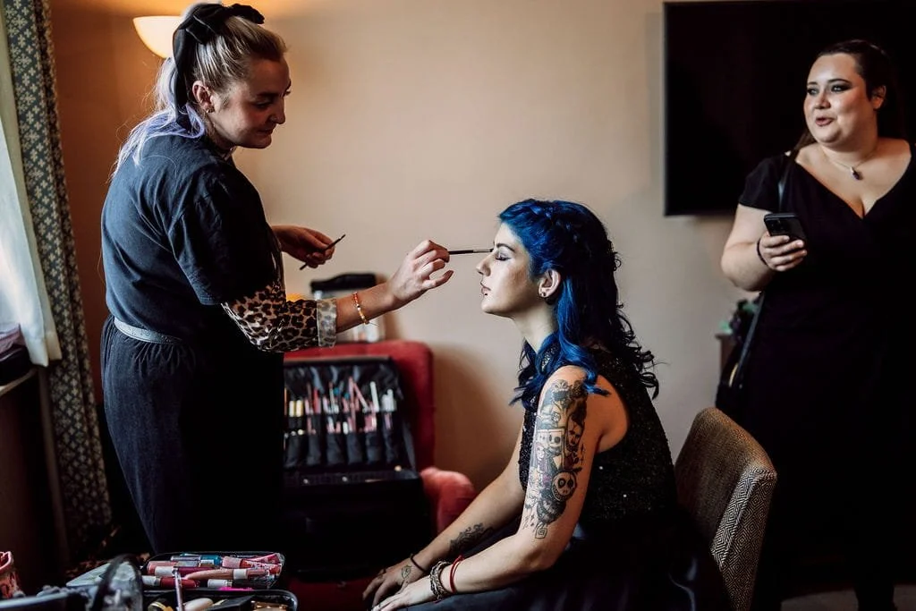 A woman with blue hair and tattoos is getting her makeup done by a makeup artist, while another woman stands nearby holding a phone.