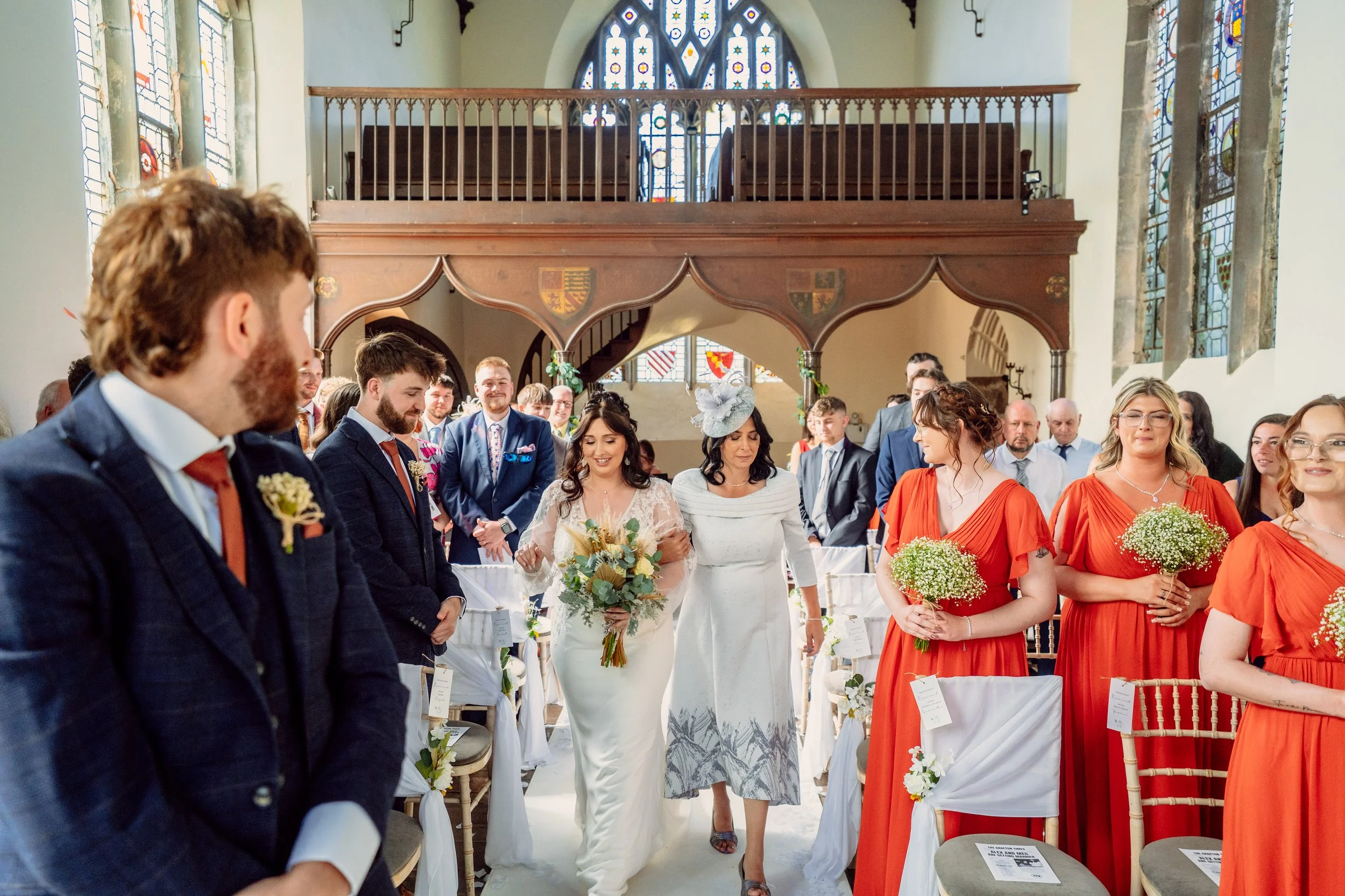A bride walking down the aisle with an older woman, likely her mother, inside a church decorated for a wedding. The bride is holding a bouquet of flowers, and the other woman is wearing a hat. Guests, including men in suits and women in orange dresse
