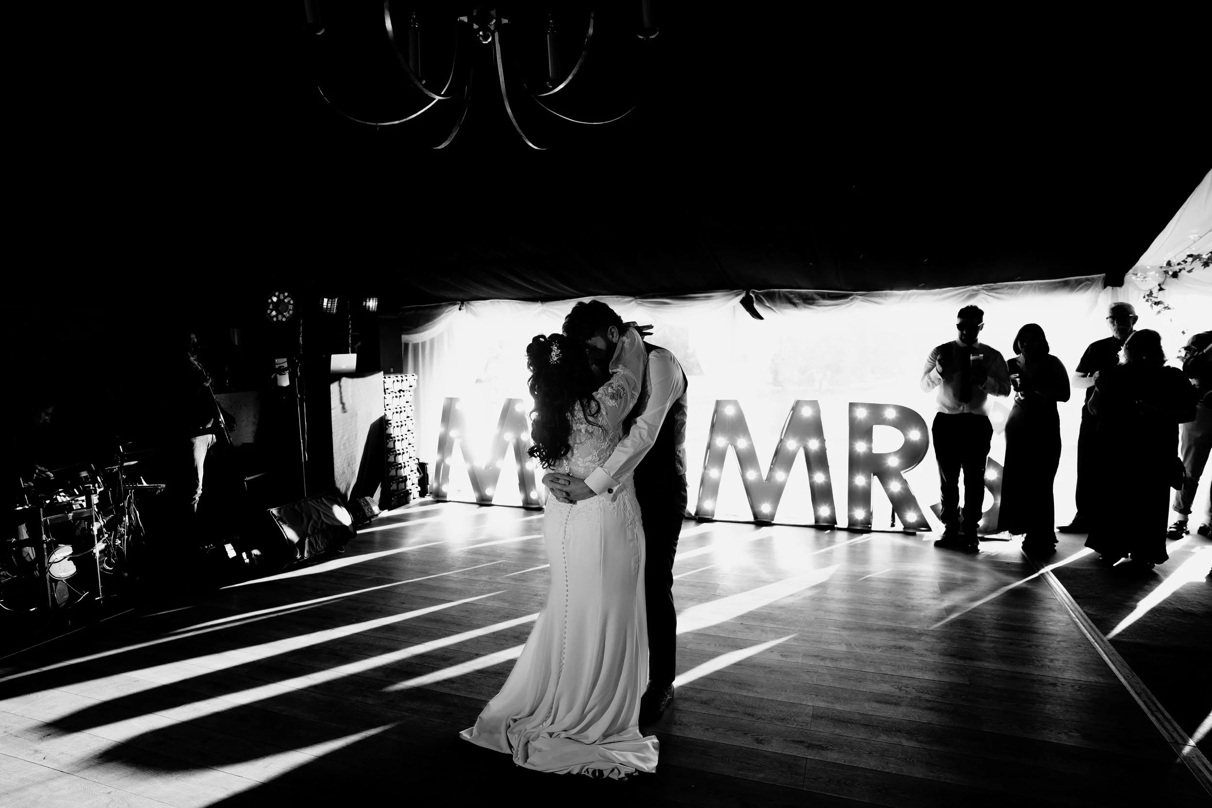 A bride and groom share a dance in a wedding reception hall, with large illuminated letters spelling 'MARRIAGE' in the background and guests watching.