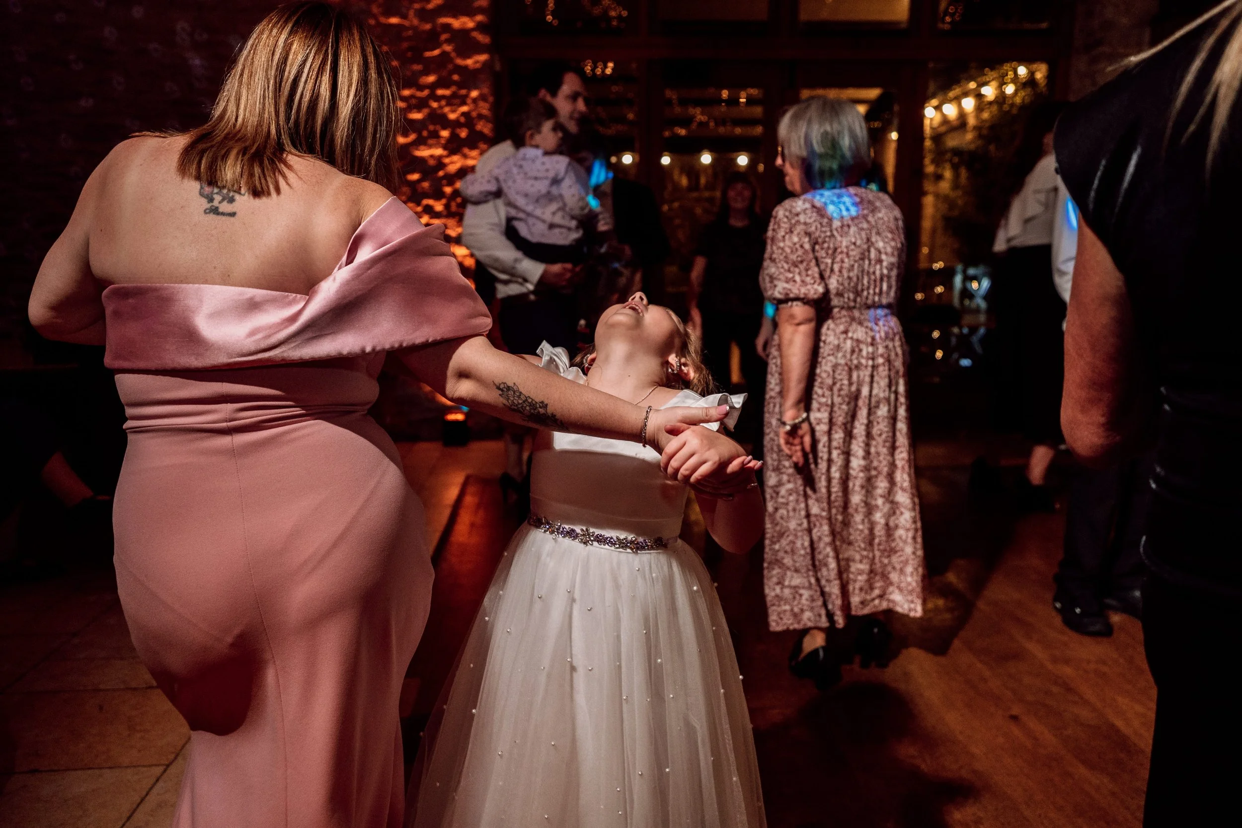 A young girl in a white dress with a sparkly belt laughing joyfully as she dances while being twirled by a woman in a pink off-shoulder gown at a festive party or celebration, with other guests in the background.