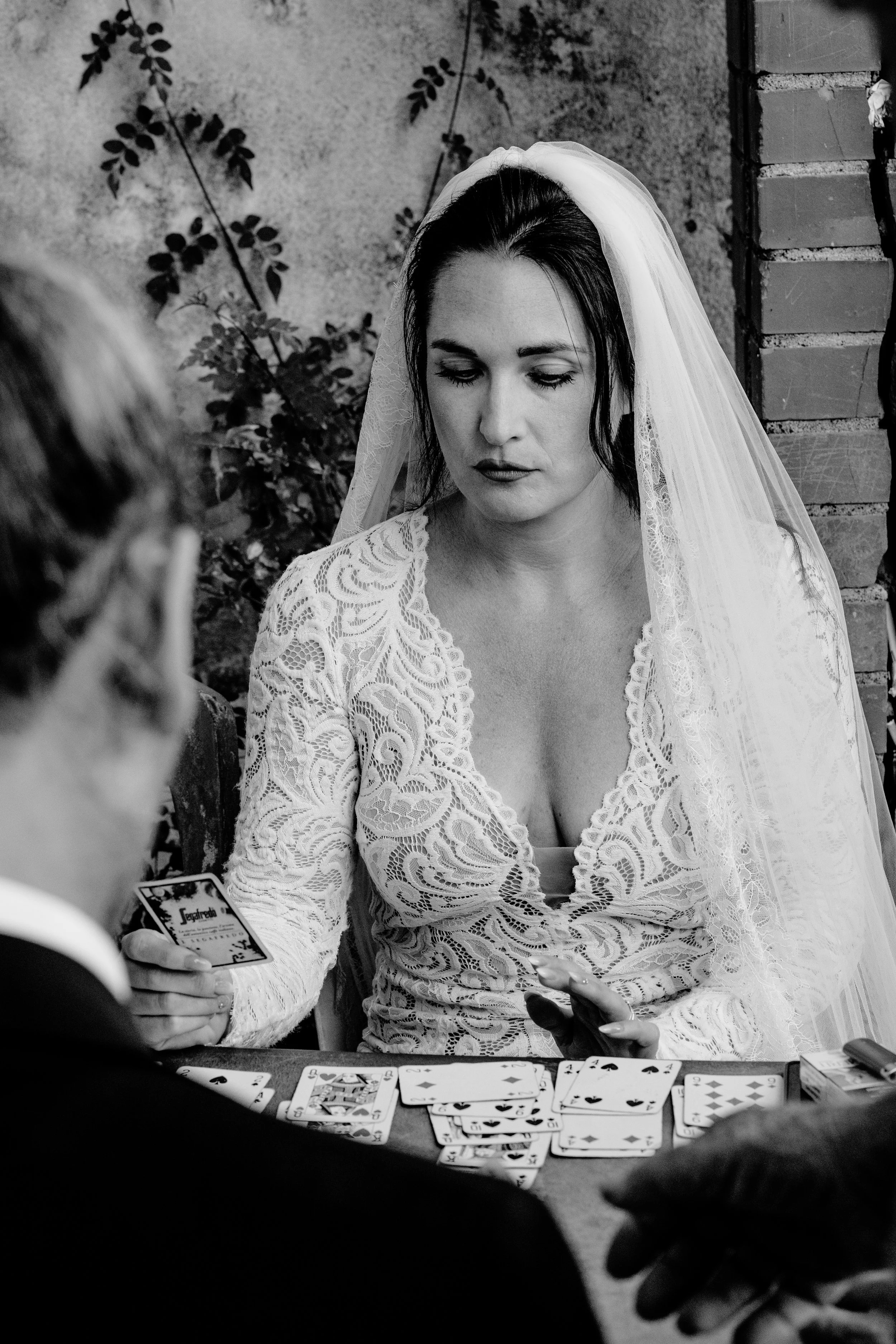 A woman in a wedding dress and veil plays cards at a table, with two other people partially visible, outdoors near a brick wall and plant.