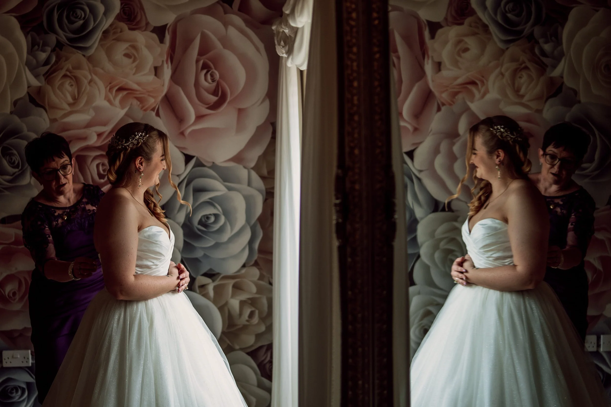 A woman in a white wedding dress is looking at herself in a mirror, with another woman assisting her, against a backdrop of large roses. The woman has long, wavy hair and is wearing jewelry.