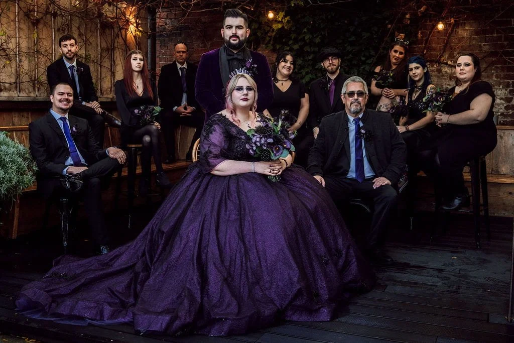 Group of people dressed in formal attire, posing outdoors with a woman in a large purple gown and man in a dark suit with glasses, in a rustic setting with a brick wall and string lights.