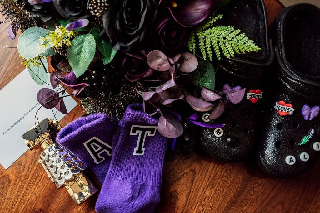 Purple wristbands, a floral arrangement with purple and green leaves, and decorated black Crocs shoes with pins and charms on a wooden surface.