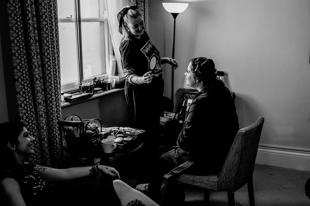 Three women in a room, one standing applying makeup to another sitting by a window, another woman sitting on a couch smiling. Makeup products are on a table.