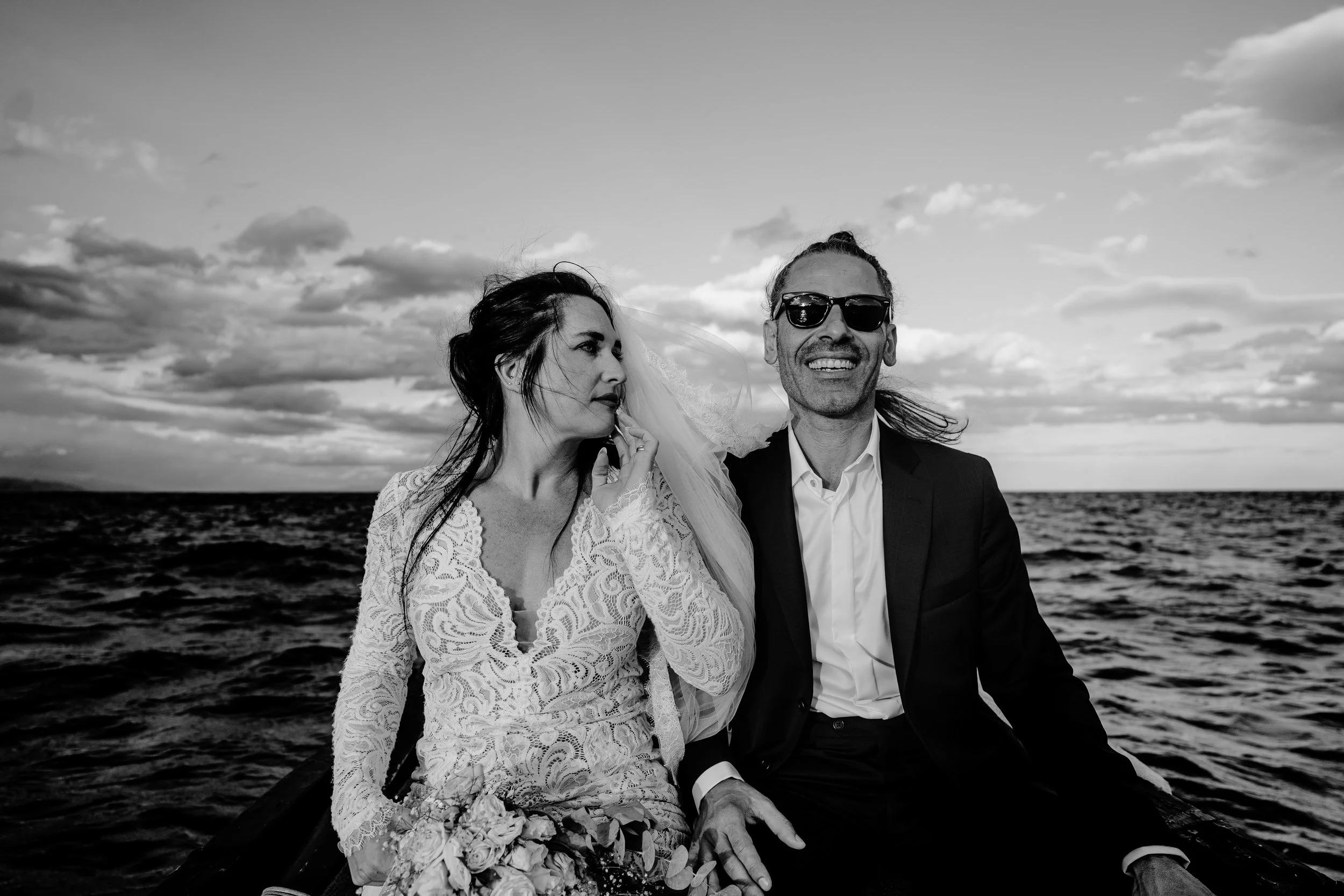 Black and white photo of a smiling man in sunglasses and a woman in a lace dress, sitting on a boat by the water during sunset.
