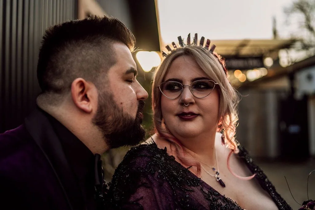 Two people facing each other outdoors during sunset, one with short dark hair and beard, the other with glasses, pink and blonde hair, and wearing a crown or tiara.