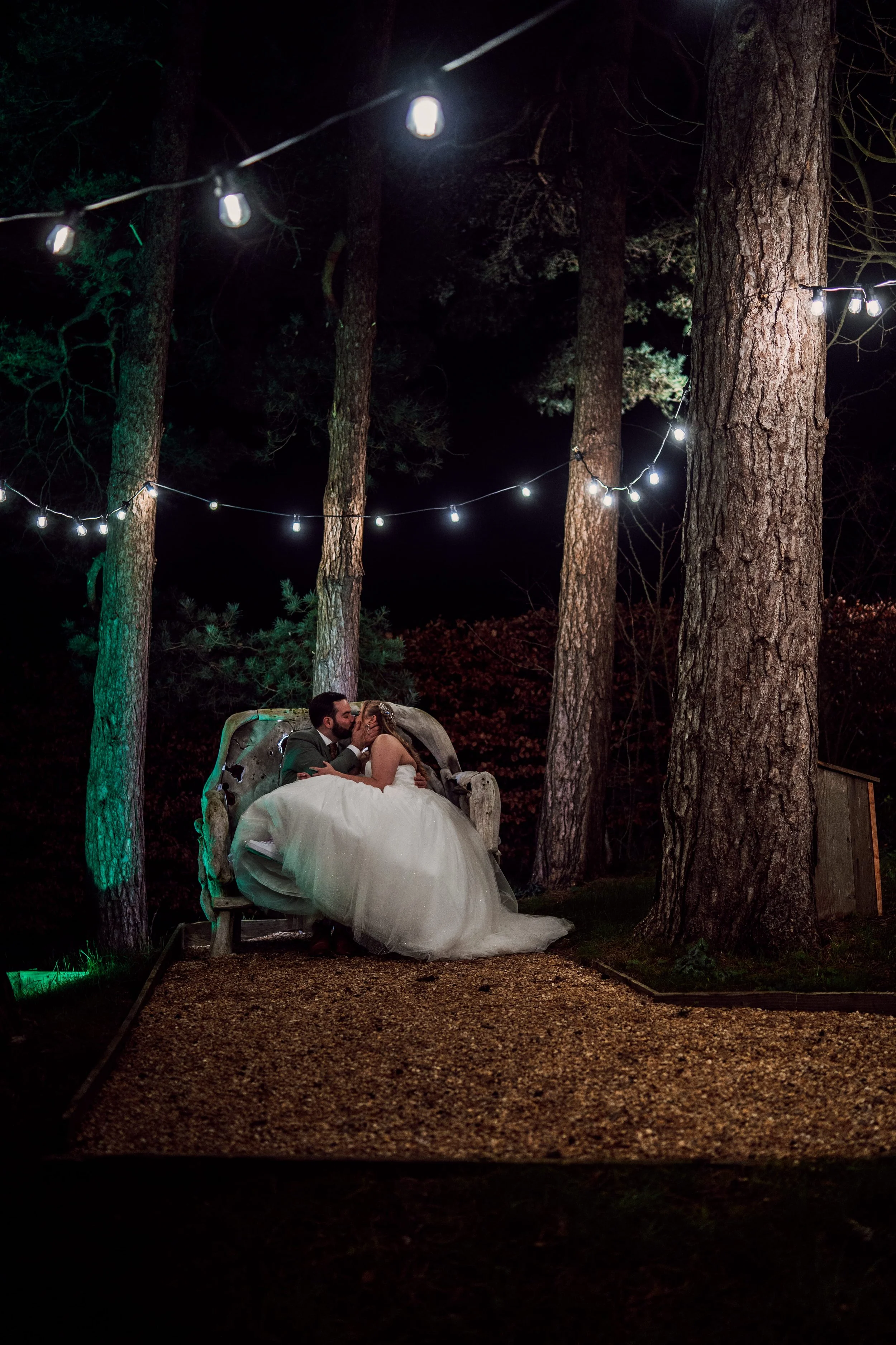 A bride and groom sitting on a wooden bench in a wooded outdoor area at night, illuminated by string lights overhead, sharing a kiss.