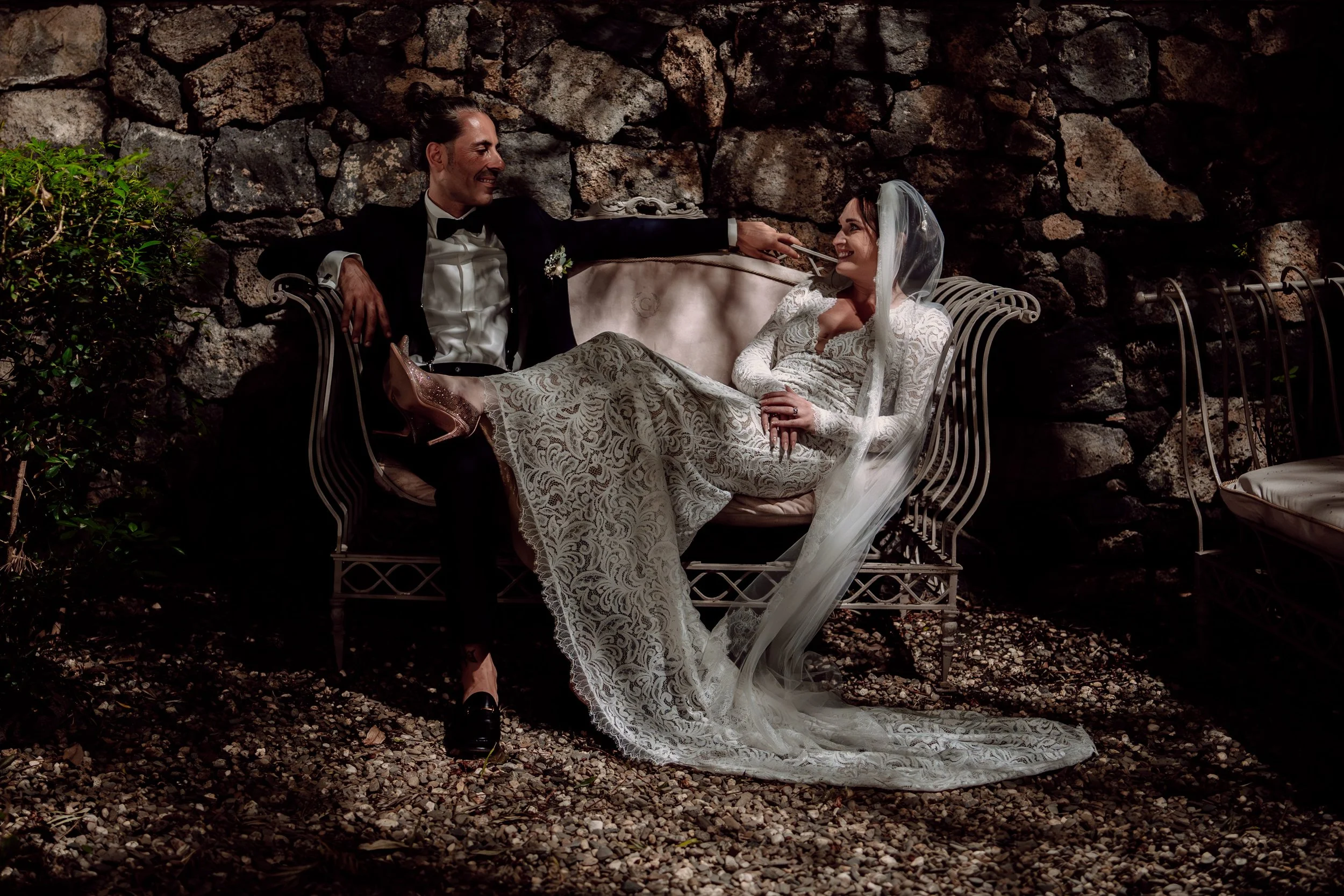 Relaxed wedding couple portrait in Sicily, seated on vintage iron garden furniture against ancient stone wall, bride in long sleeve lace gown with flowing veil, dramatic shadow and light - destination wedding photography by Khya Watts