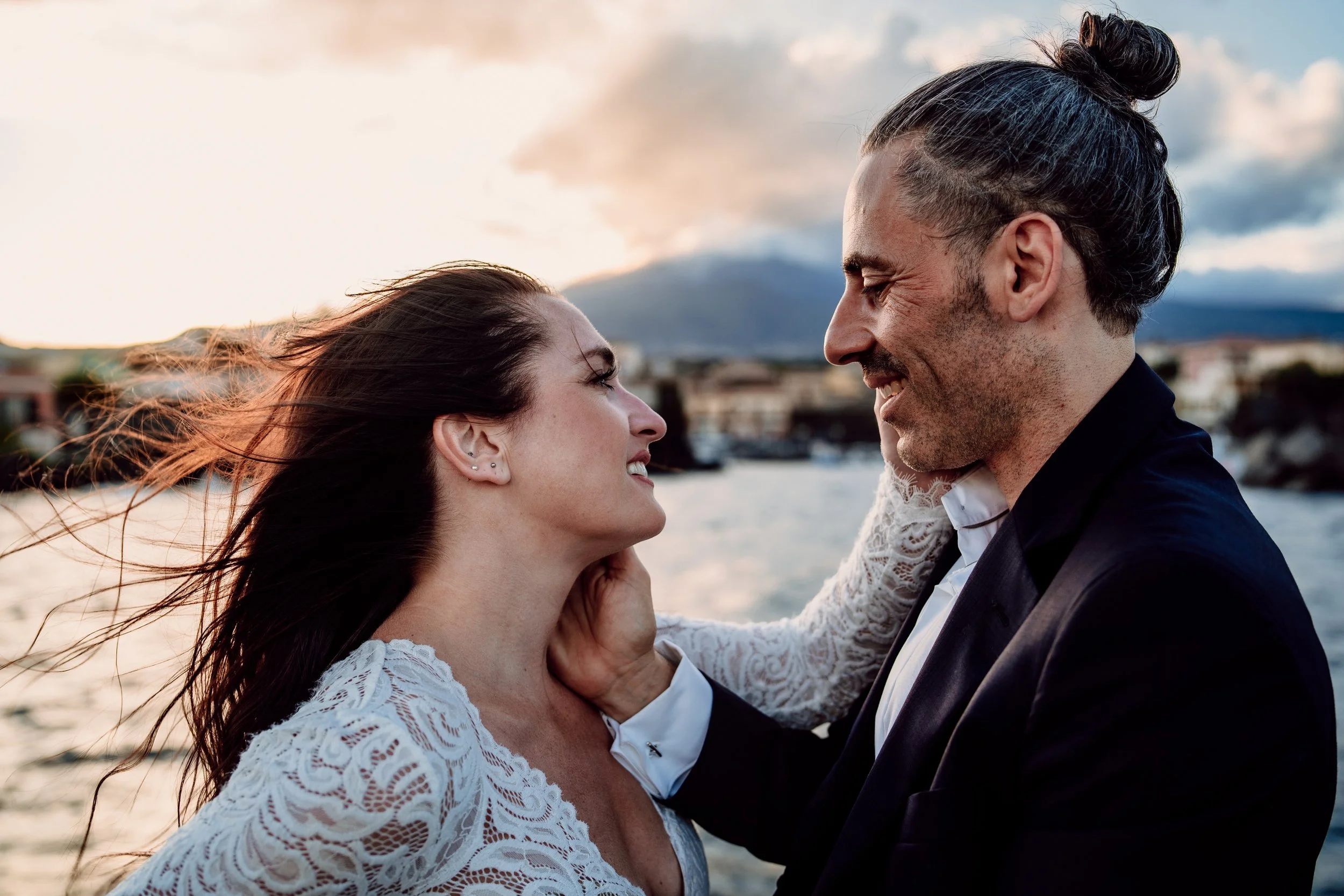 A man and woman are smiling at each other outdoors near a body of water, with hills and buildings in the background, during sunset.