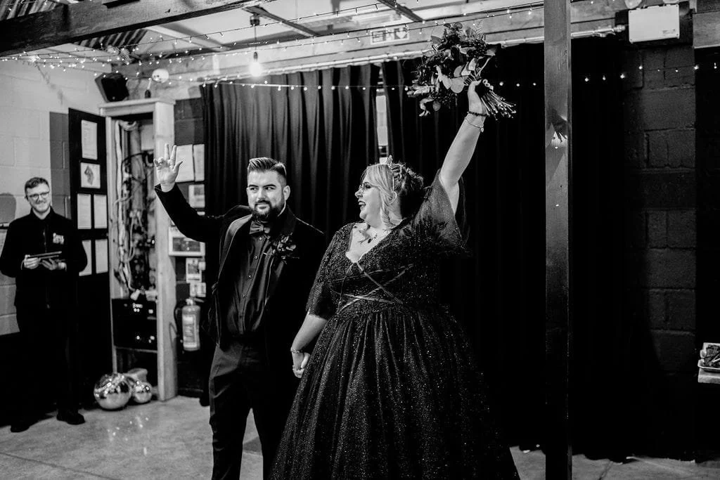 A woman in a sparkling black dress holds a bouquet high while a man in a suit waves and looks on during a celebration in an indoor venue with string lights.