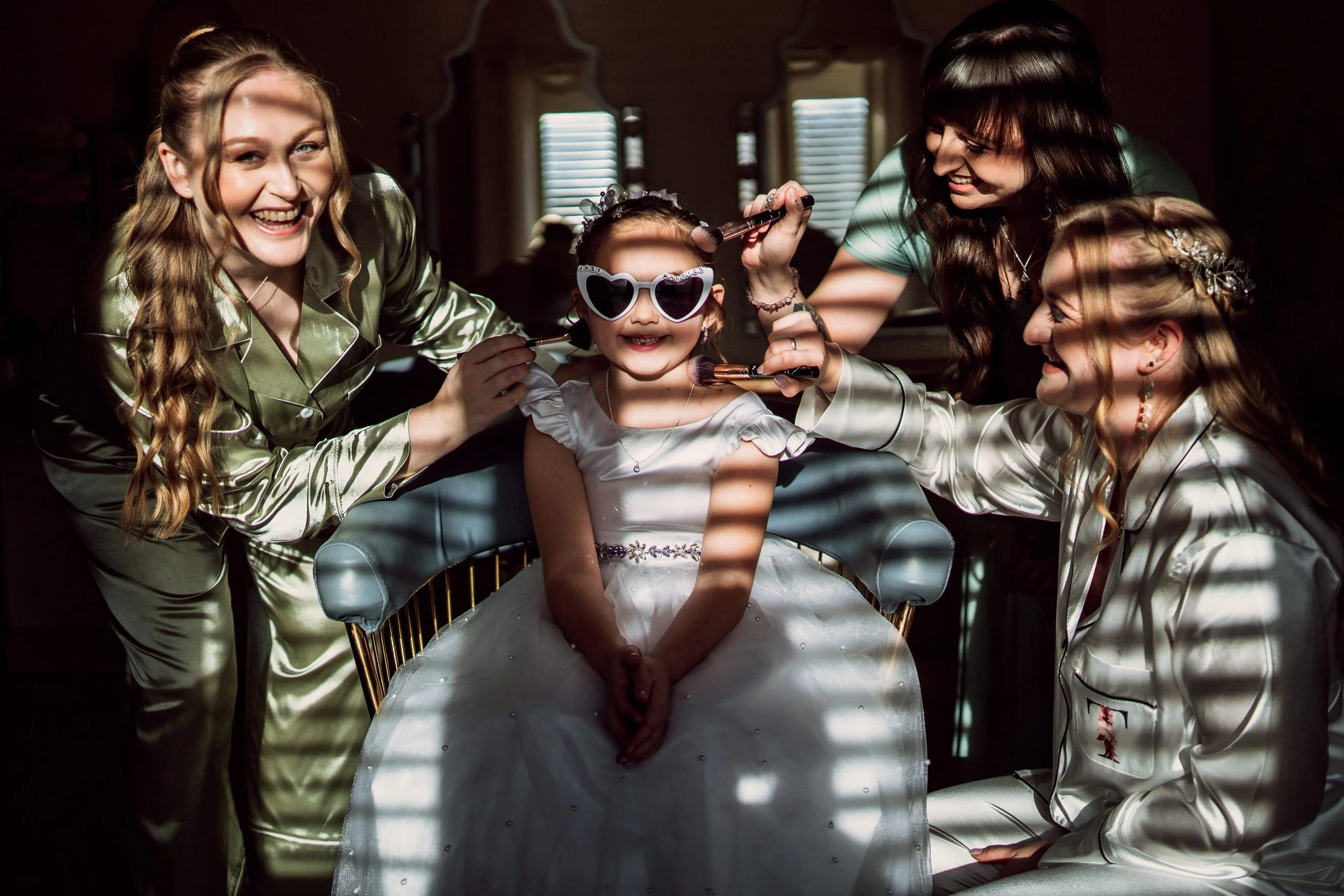 A young girl in a white dress and heart-shaped sunglasses sitting in a chair, surrounded by four women in satin pajamas applying makeup and smiling, with sunlight casting stripes through window blinds.