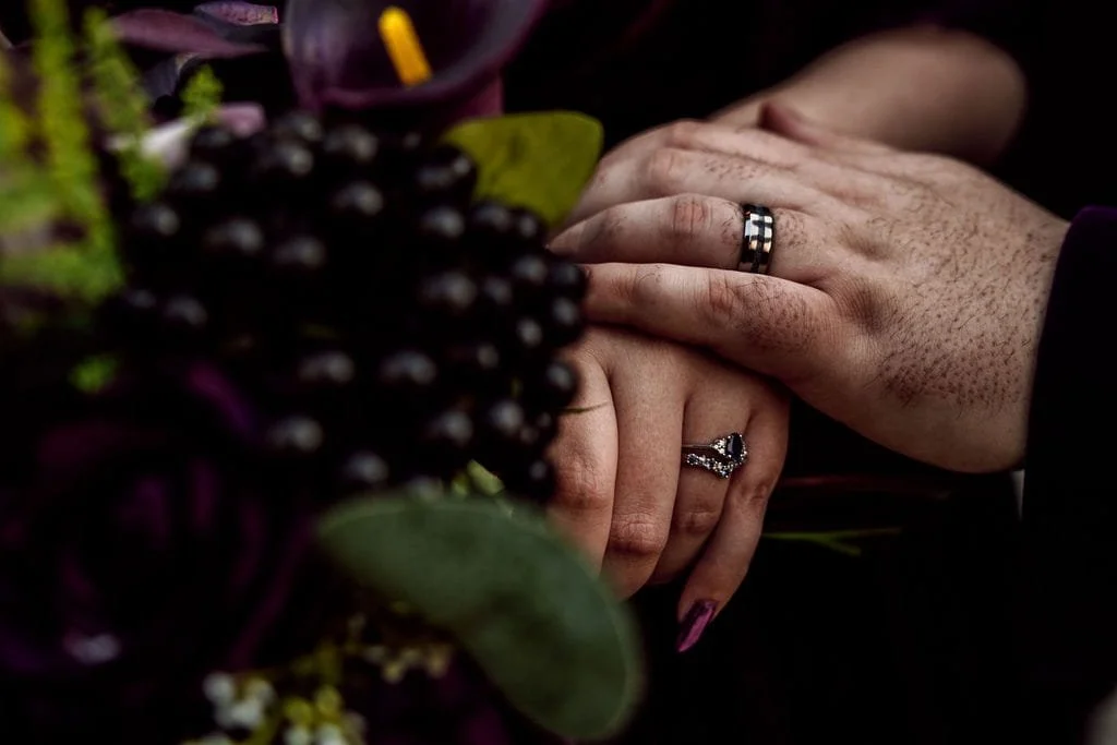 Close-up of two hands with wedding and engagement rings touching among dark flowers and greenery.