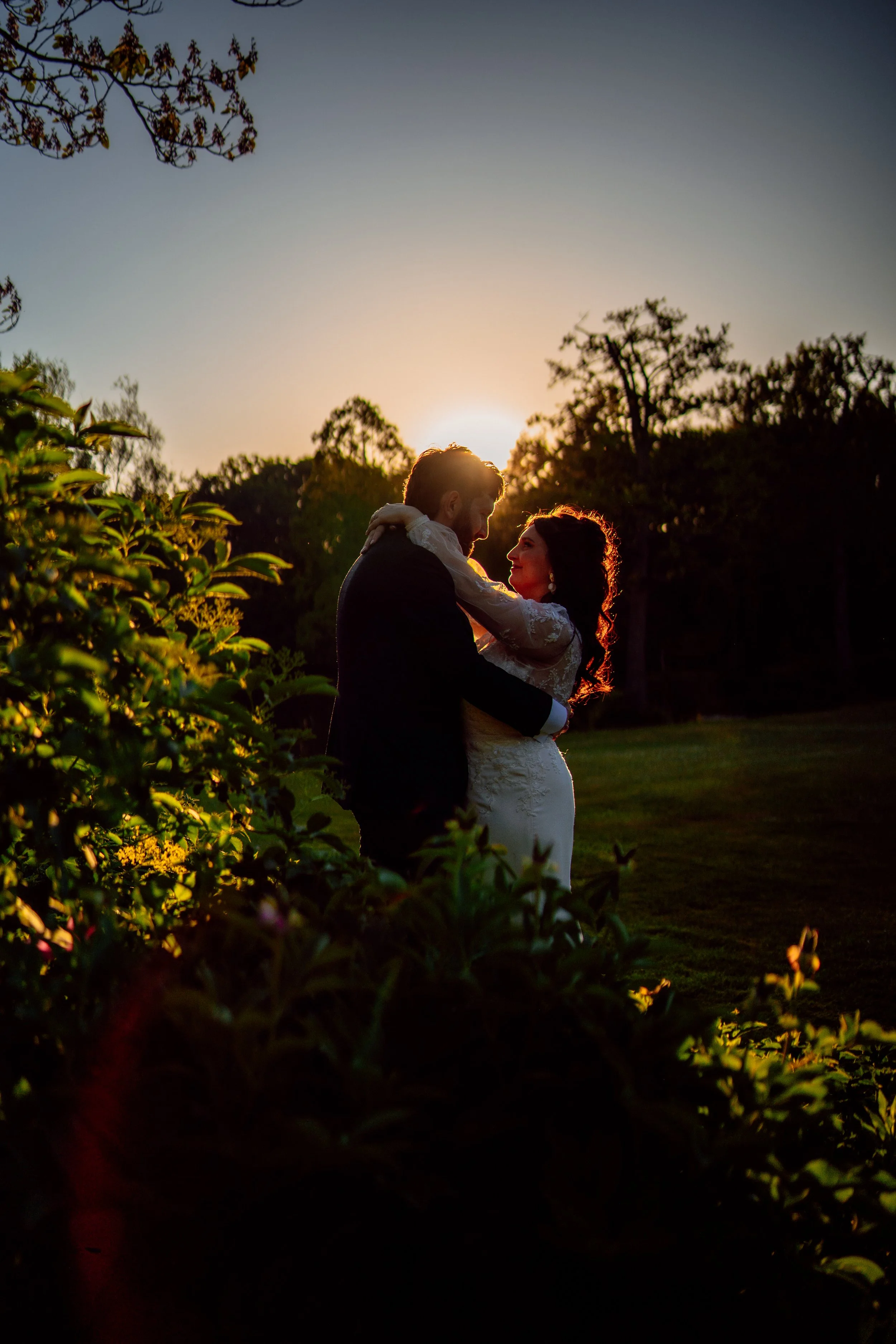 Silhouette of a newlywed couple embracing outdoors at sunset, surrounded by greenery and trees, with the sun setting behind them.