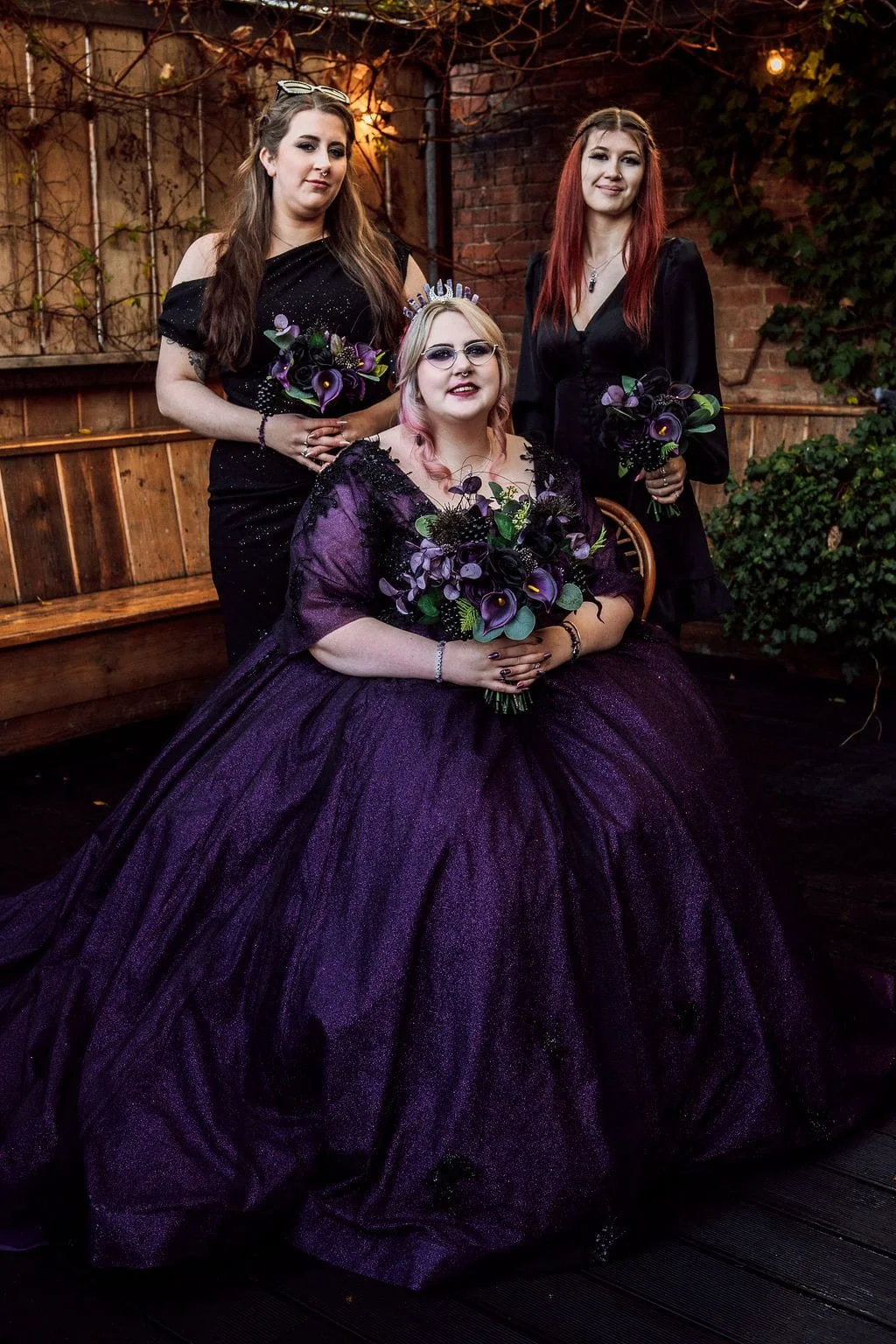 Three women in formal dresses holding purple flower bouquets outdoors, with a brick wall and vines in the background, during a twilight evening.
