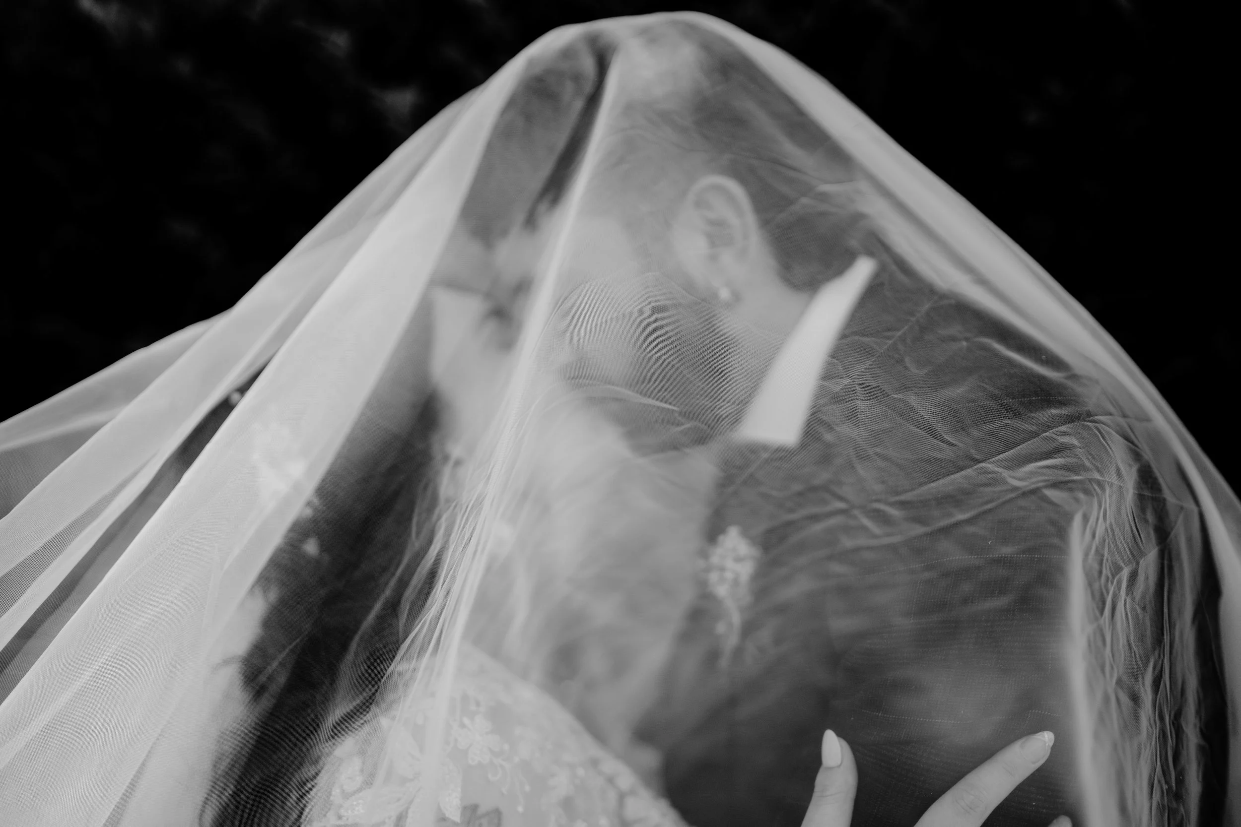 Bride and groom's hands touching, with a veil covering their faces, on their wedding day.