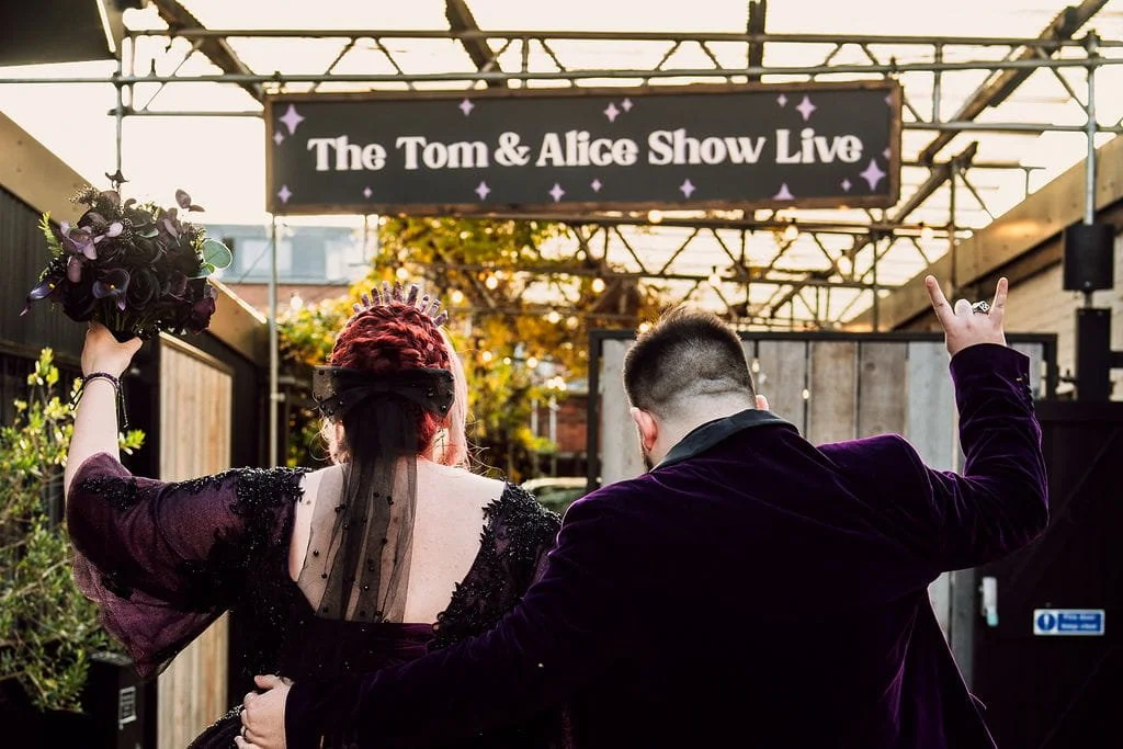 Two people walking toward an outdoor stage with a sign that reads 'The Tom & Alice Show Live.' One person has red hair and is wearing a black dress with a veil, holding a bouquet, while the other has dark hair and is wearing a purple jacket, making a