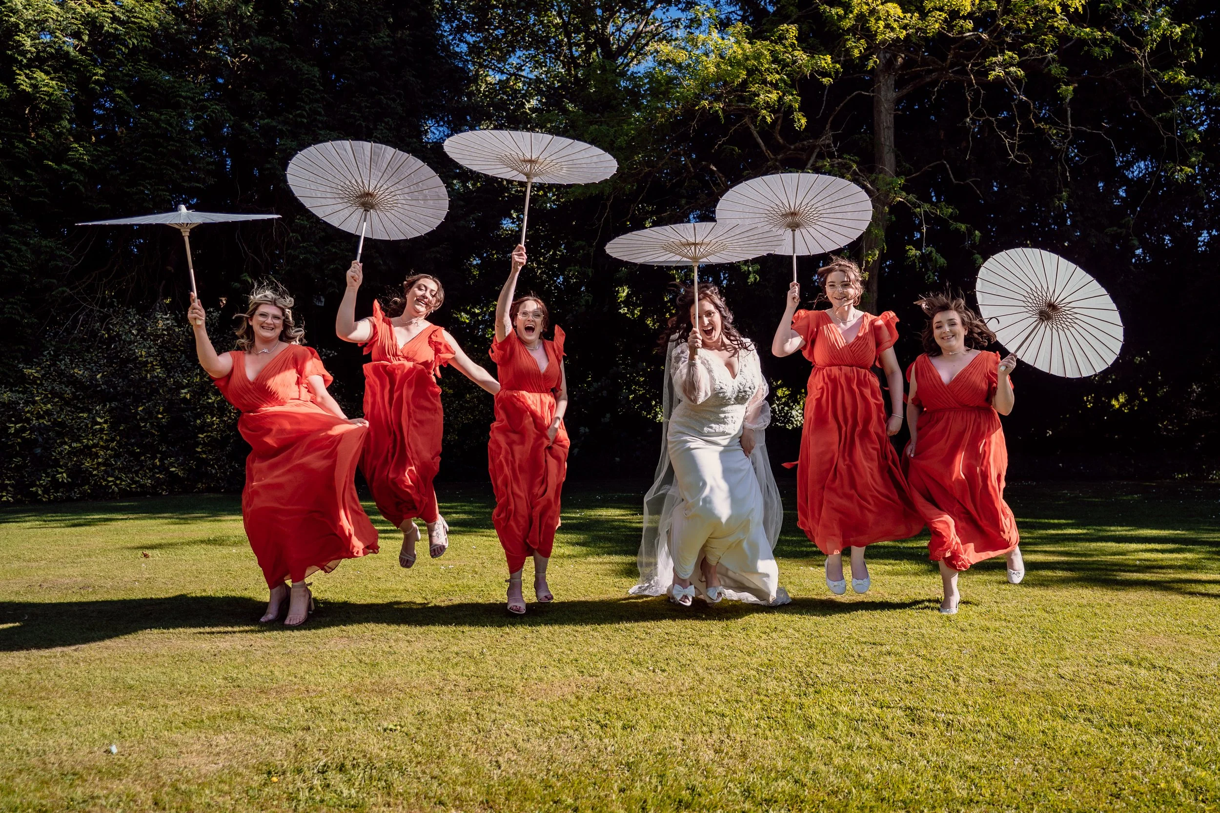 Bride and five bridesmaids in red dresses and white dress, holding white parasols, jumping on grassy lawn with trees in background.