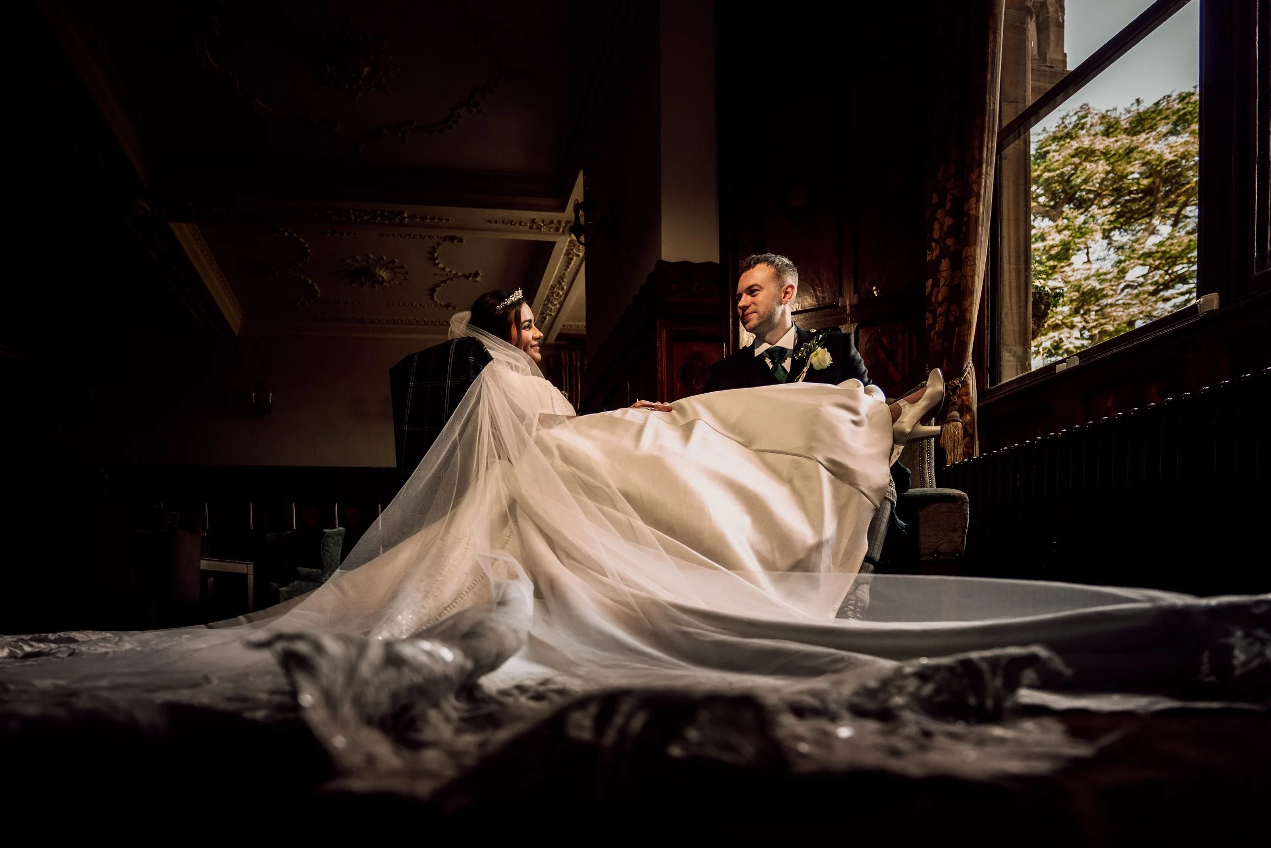 Dramatic low angle wedding portrait at Ettington Park, bride and groom seated in a dark ornate room, cathedral length veil pooling across the floor, moody natural light from tall gothic windows - alternative wedding photography by Khya Watts