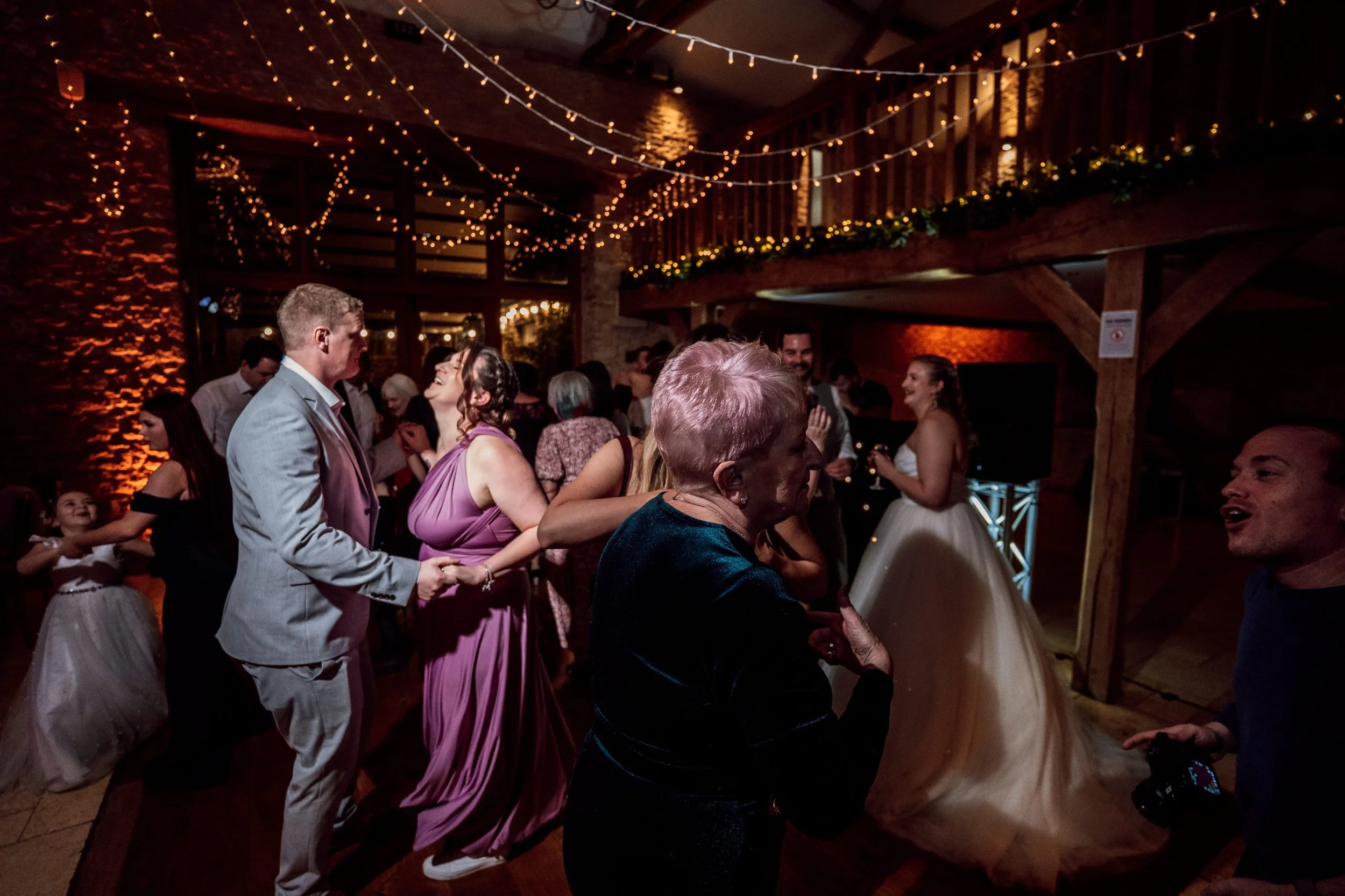 People dancing and celebrating at a wedding reception in a decorated venue with string lights and wooden accents.