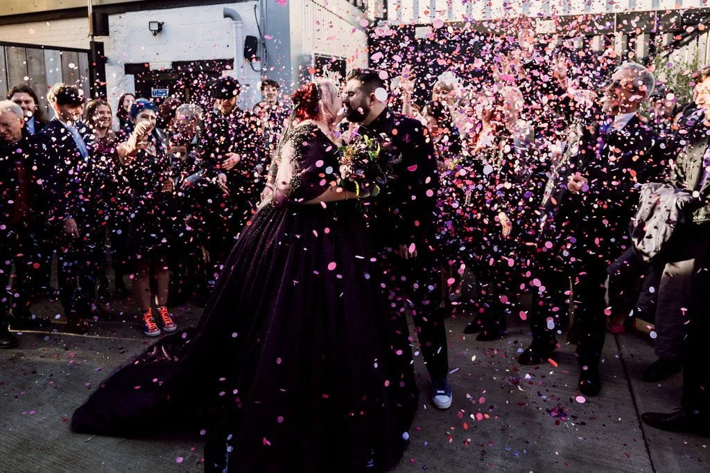 Couple getting married outdoors, surrounded by guests throwing pink and purple confetti, with celebratory atmosphere.