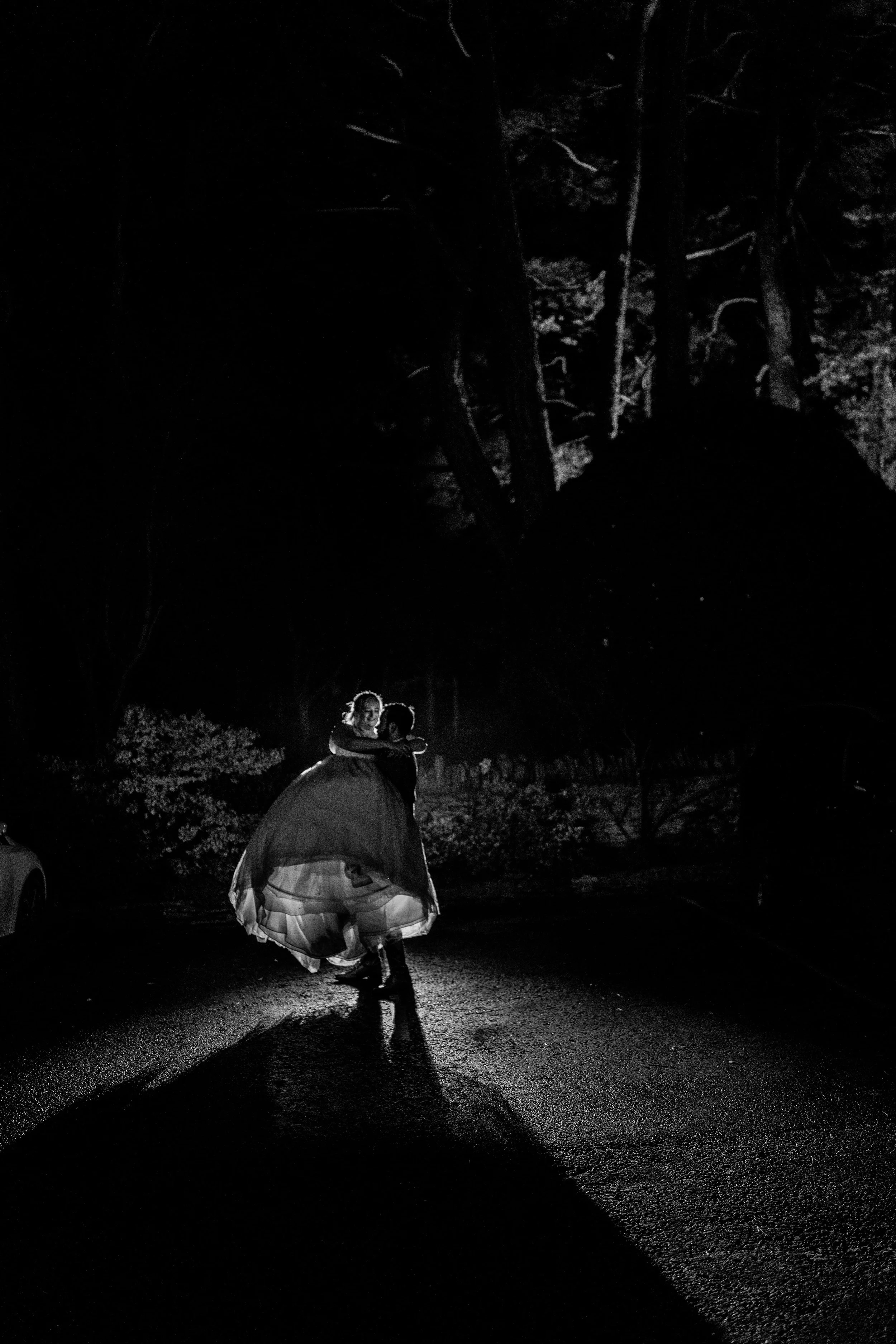 A black and white photo of a couple dancing in the street at night, illuminated by a single light source, with trees and cars in the background.