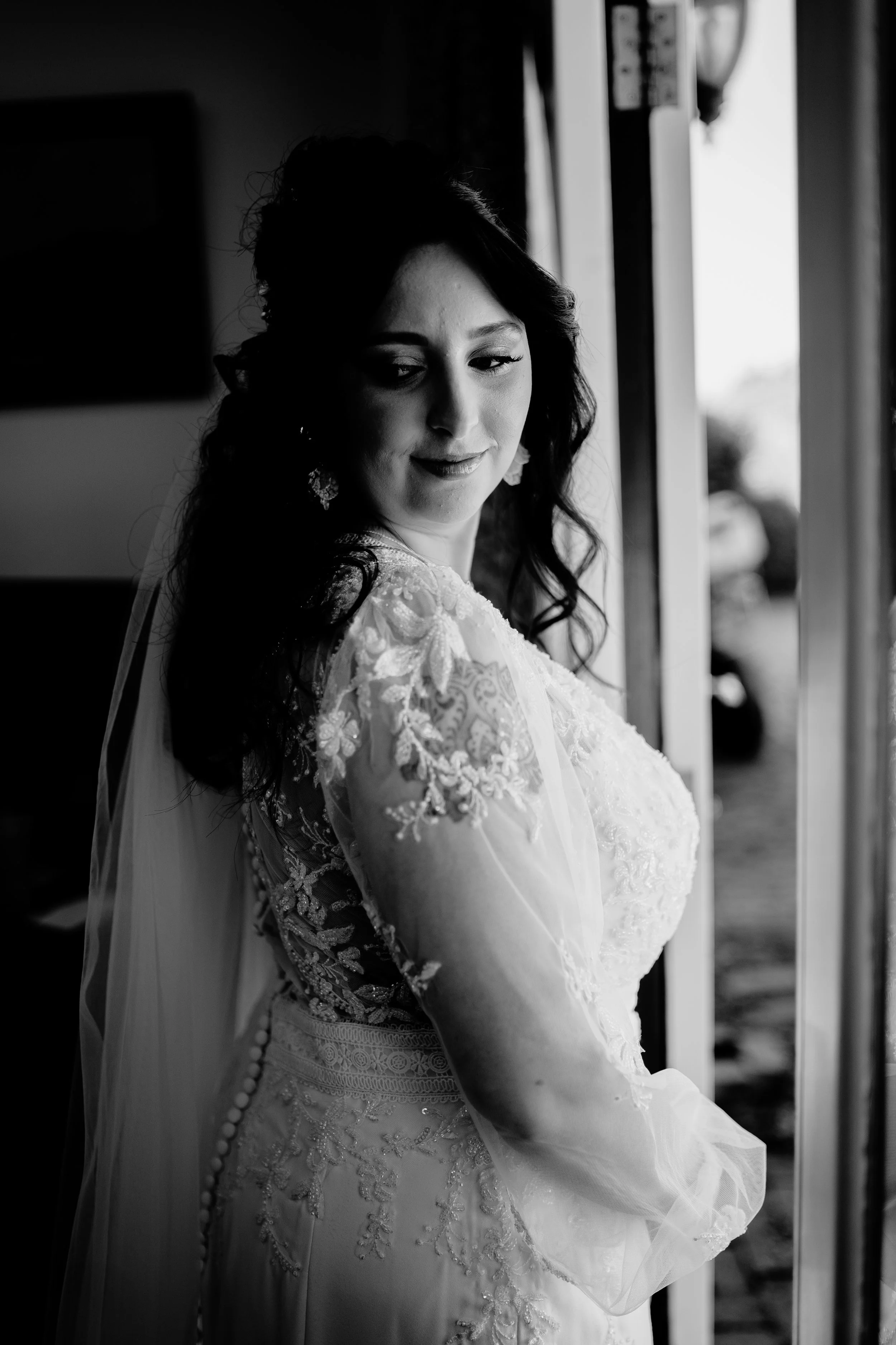 A woman in a lace wedding dress standing by a window, looking down with a gentle smile, in black and white