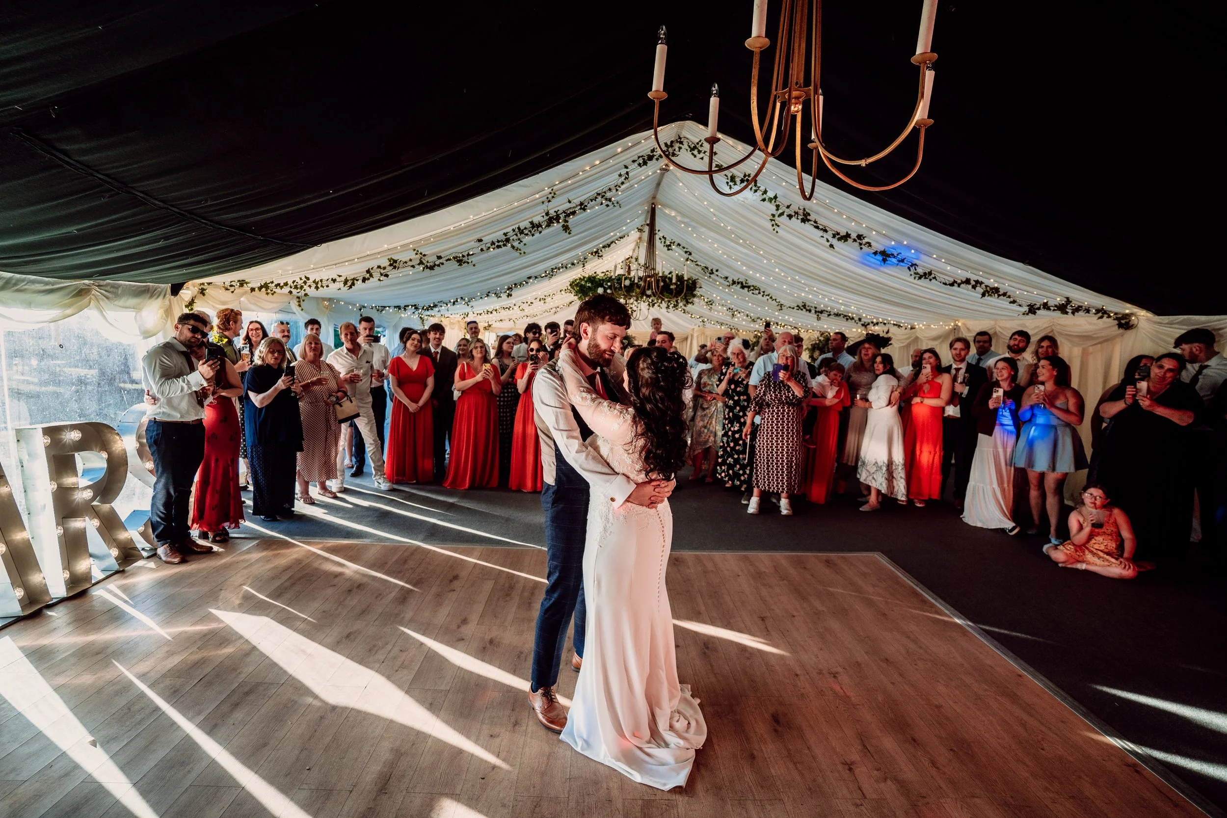 A bride and groom sharing their first dance at a wedding reception under a decorated tent with string lights and greenery, with guests watching and taking photos.