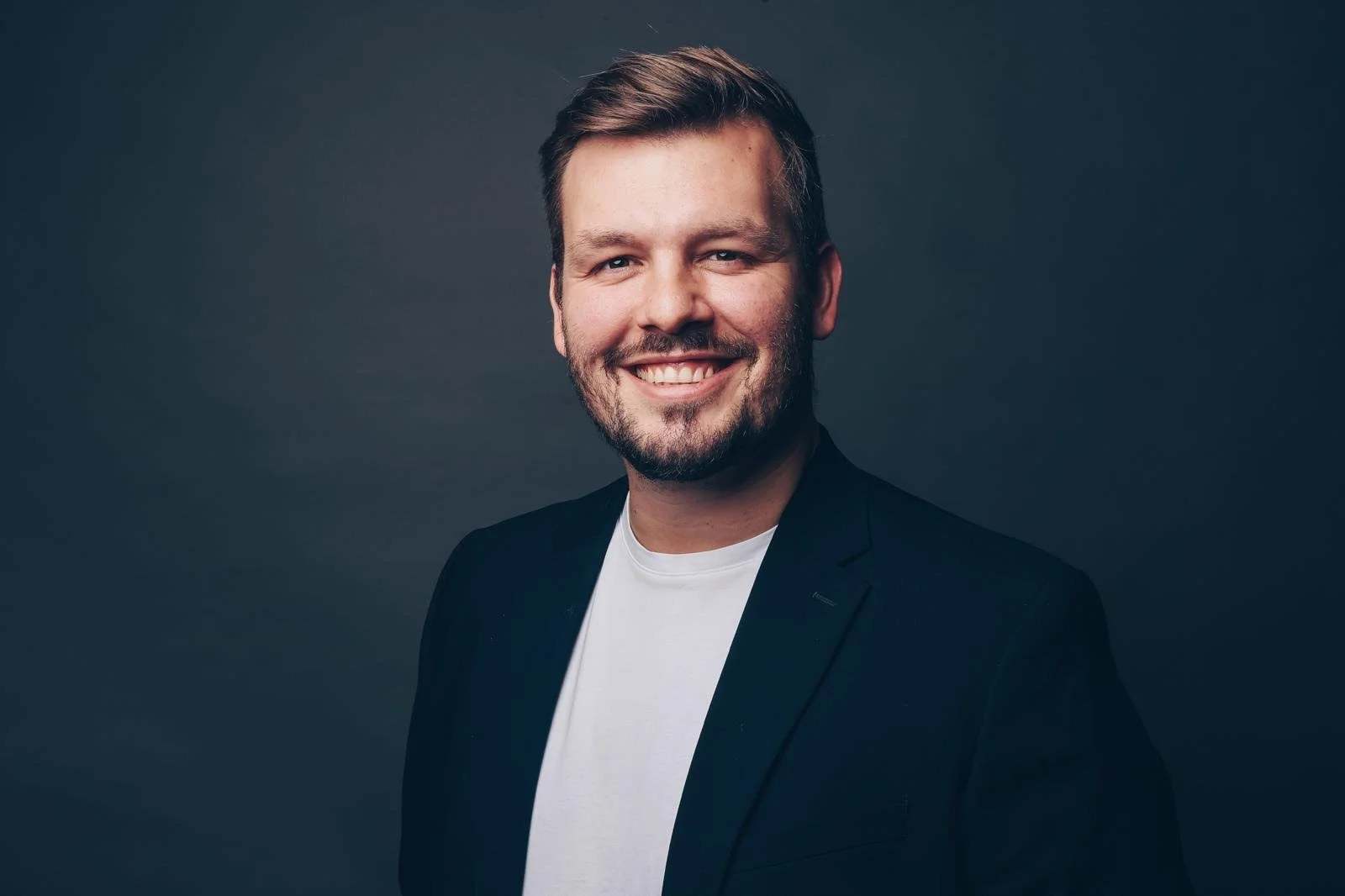Lächelnder Mann mit dunklem Sakko und weißem T-Shirt vor dunklem Hintergrund.