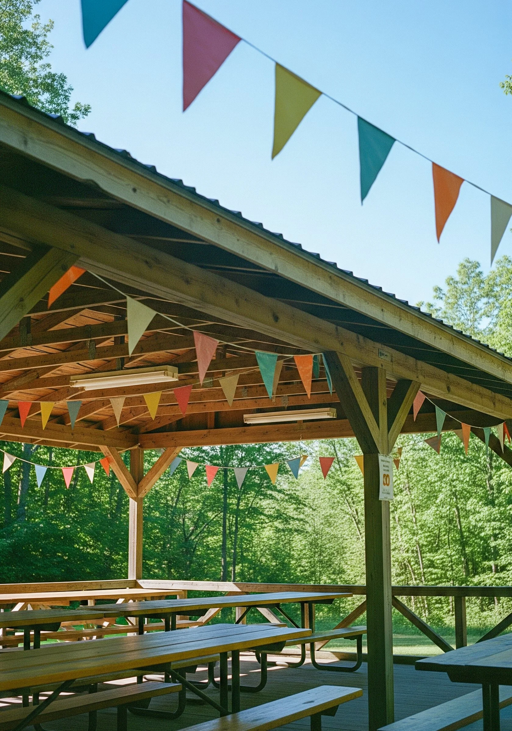 Ein outdoor Veranstaltungsbereich mit Holztischen und Bänken, bunt geflebte Wimpel als Dekoration, umgeben von grünen Bäumen bei Tageslicht.