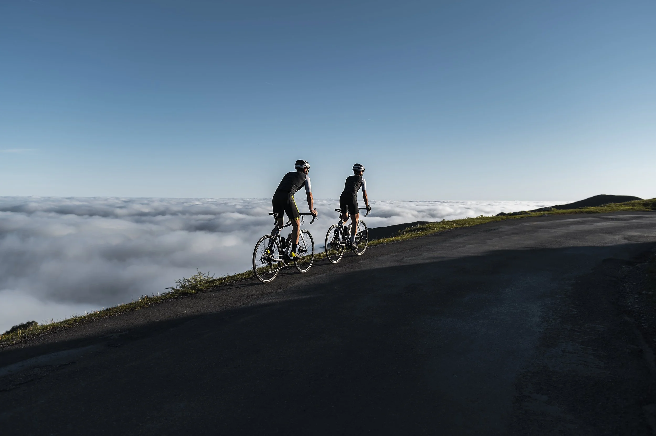 Ein Sportler und eine Sportlerin fahren an einem sonnigen Morgen gemeinsam Fahrrad auf einer Bergstraße, die von nebelverhangenen Bergen umgeben ist