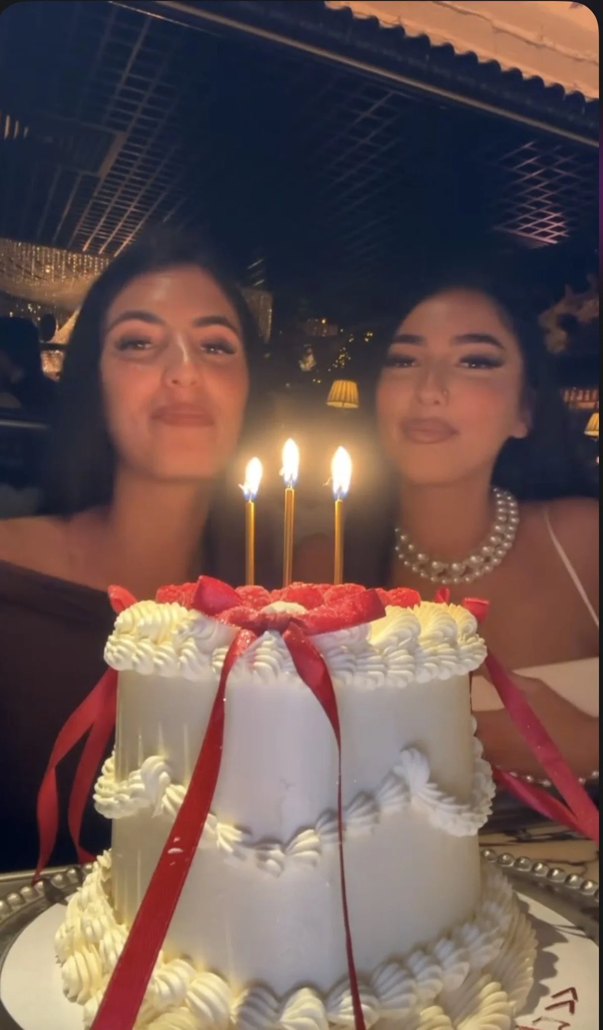 Two women celebrating birthday with a cake that has three lit candles, strawberries, and red ribbons.