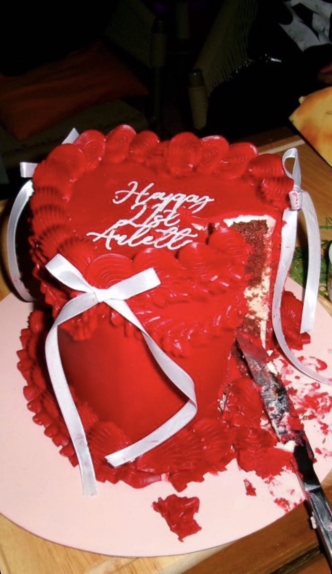 A heart-shaped red cake with red decorative shells and white ribbons, partially cut, with a message that reads 'Happy List of Aulcie.'