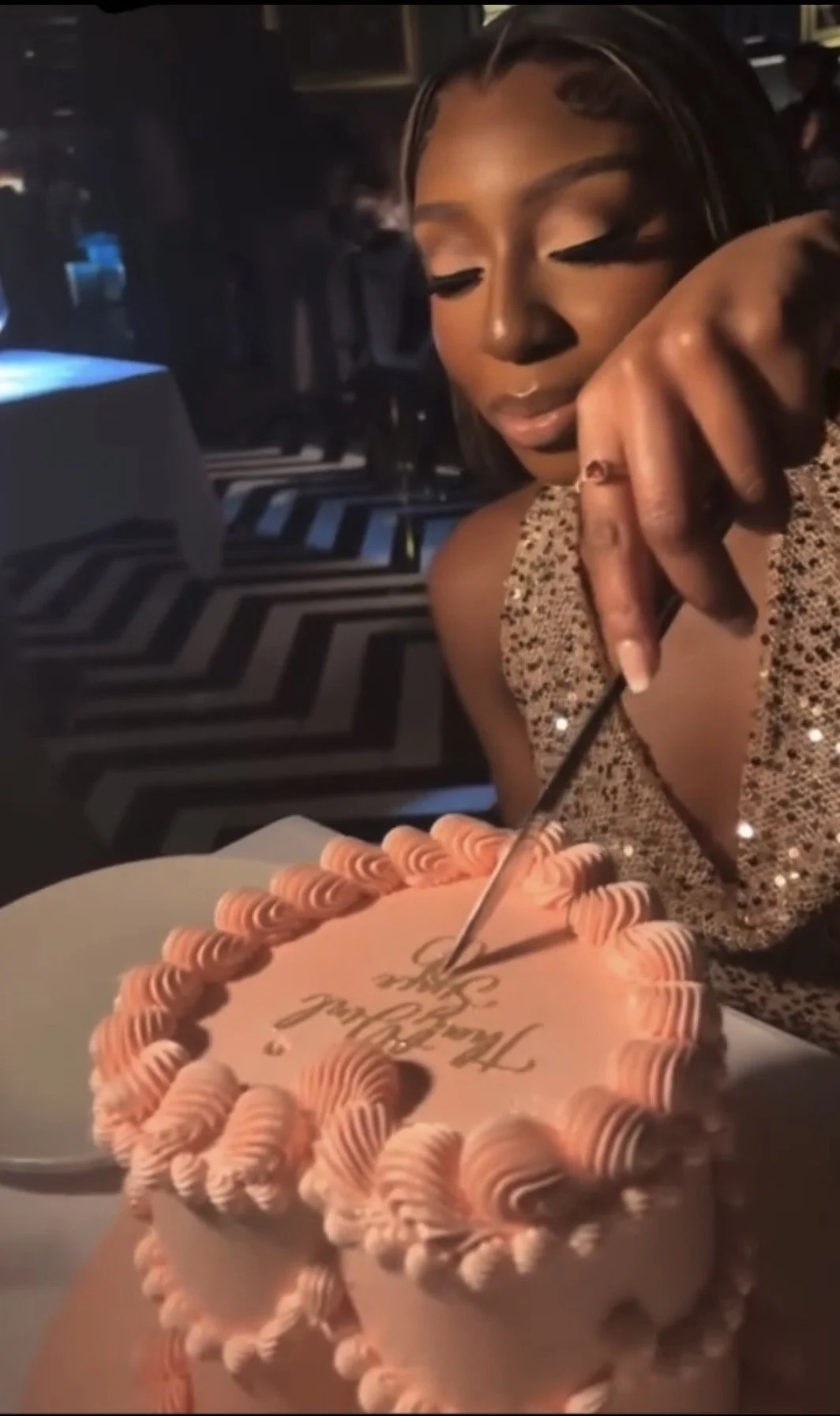 A woman with makeup and earrings is at a birthday celebration, holding a knife to cut a pink birthday cake with orange border decorations, inscribed with 'Happy Birthday.'