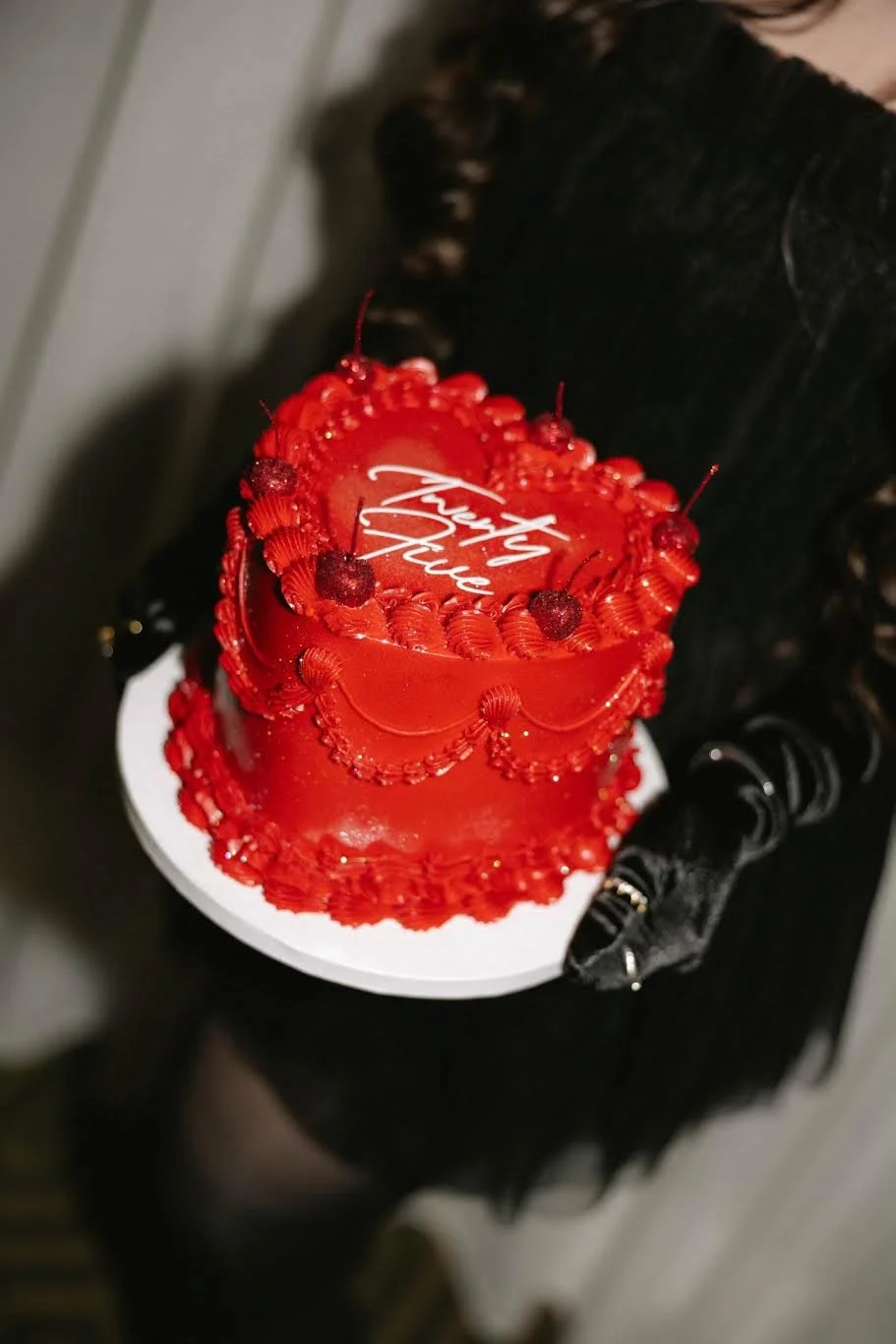 A person in black clothing holding a red, heart-shaped birthday cake decorated with red icing, strawberries, and gold accents, with the words "Happy Birthday" written on top.