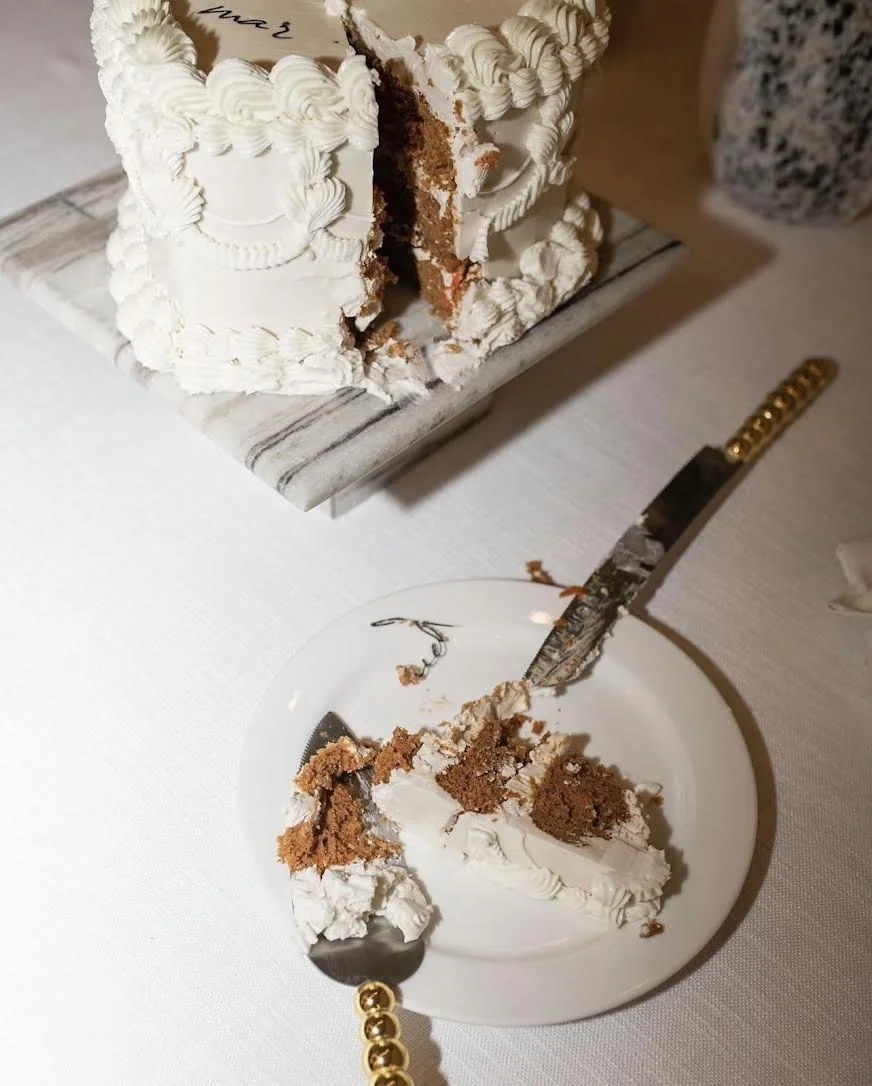 A partially sliced layered cake with white frosting and piped decorations on a marble cake board, with some cake on a white plate with a bite taken out, and a cake serving knife with a golden handle nearby on a white tablecloth.