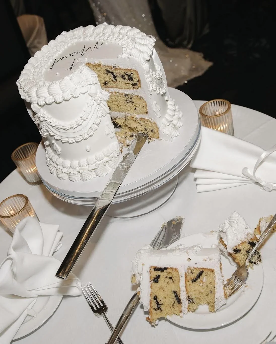 A partially sliced white wedding cake with multiple layers and chocolate chips inside, decorated with white icing and piped decorations, placed on a round white cake stand. There is a serving knife inserted into the cake, and some slices have been served onto small plates. The table setting includes white napkins, small candles in holders, and forks.