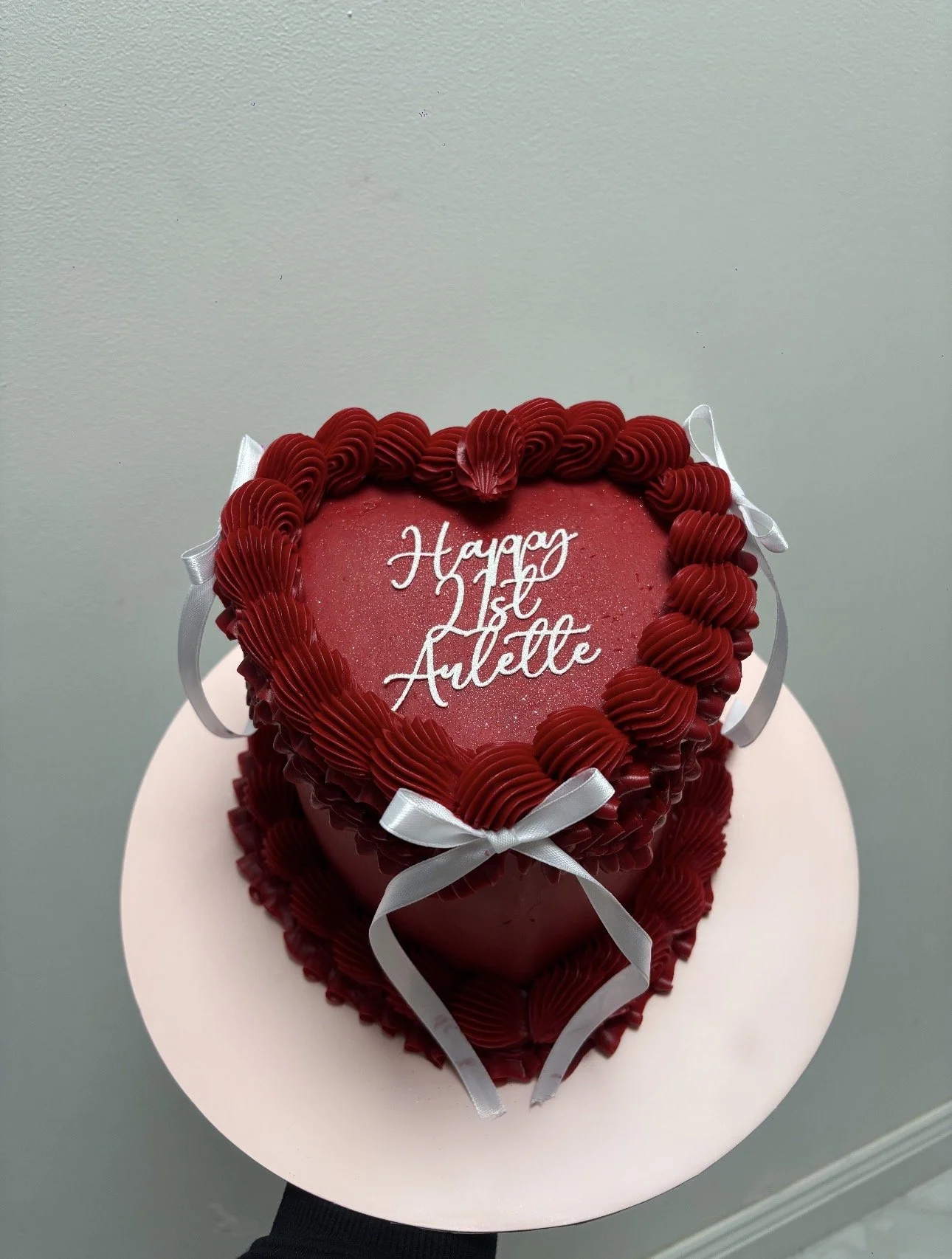 A heart-shaped red cake with decorative icing around the edges, tied with white ribbons, and a message that says 'Hayay 1st Avelle' written on top in white script.