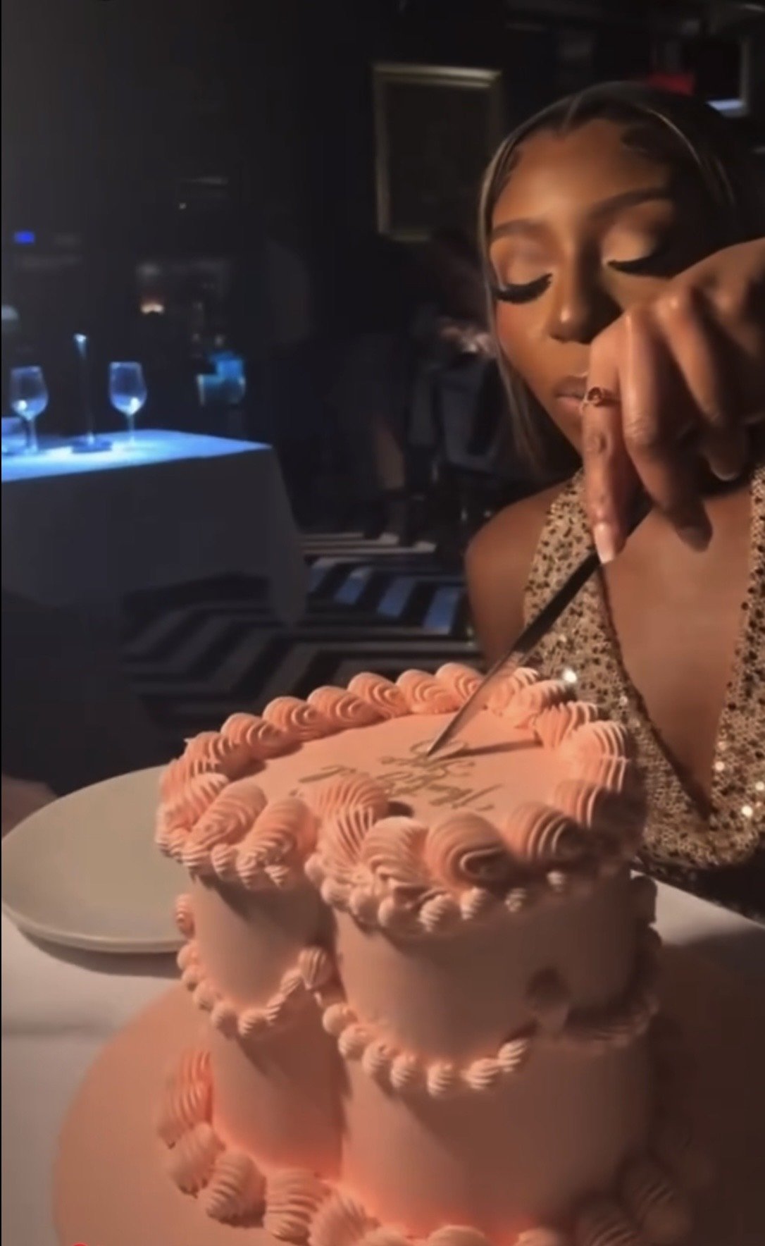 A woman in a dress with a sparkly pattern is cutting into a pink, multi-tiered birthday cake with beige and pink frosting, using a knife. There are wine glasses and a table in the background.