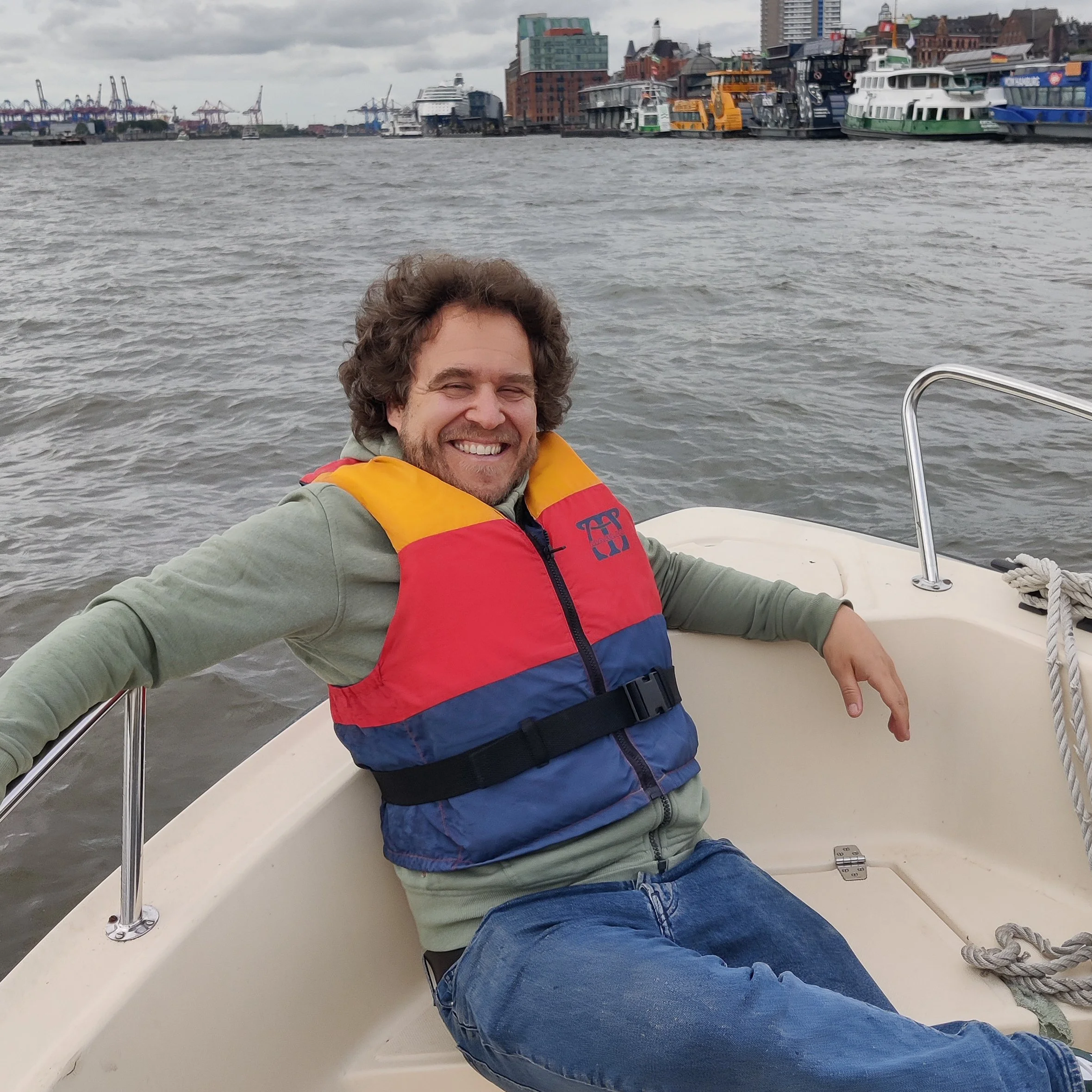 Ein lachender Mann sitzt auf einem Boot im Wasser, trägt eine rote, gelbe und blaue Schwimmweste und hat dunkle, lockige Haare. Im Hintergrund ist der Hamburger Hafen mit Gebäuden und mehreren anlegenden Booten zu sehen.