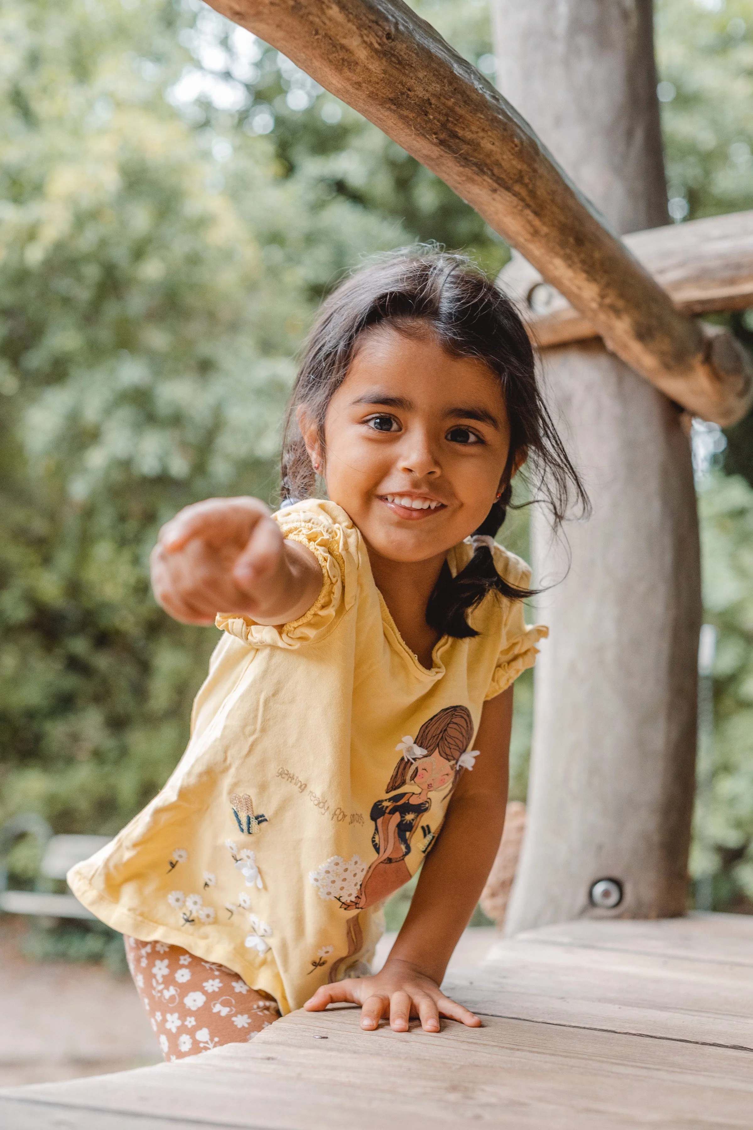 Ein lachendes Mädchen in einem gelben T-Shirt, das Richtung Kamera zeigt, während sie auf einem Klettergerüst aus Holz im Freien steht.