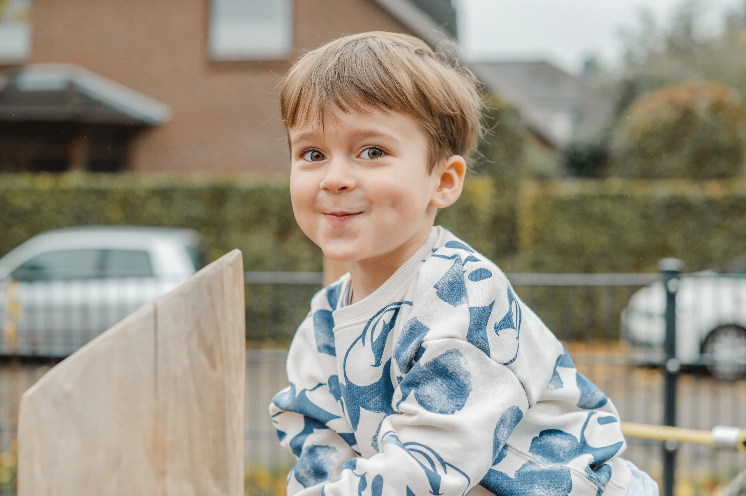 Ein lachender Junge mit braunen Haaren und blauen Augen, trägt ein weißes Sweatshirt mit blauen Mustern, spielt draußen auf einem Klettergerüst.