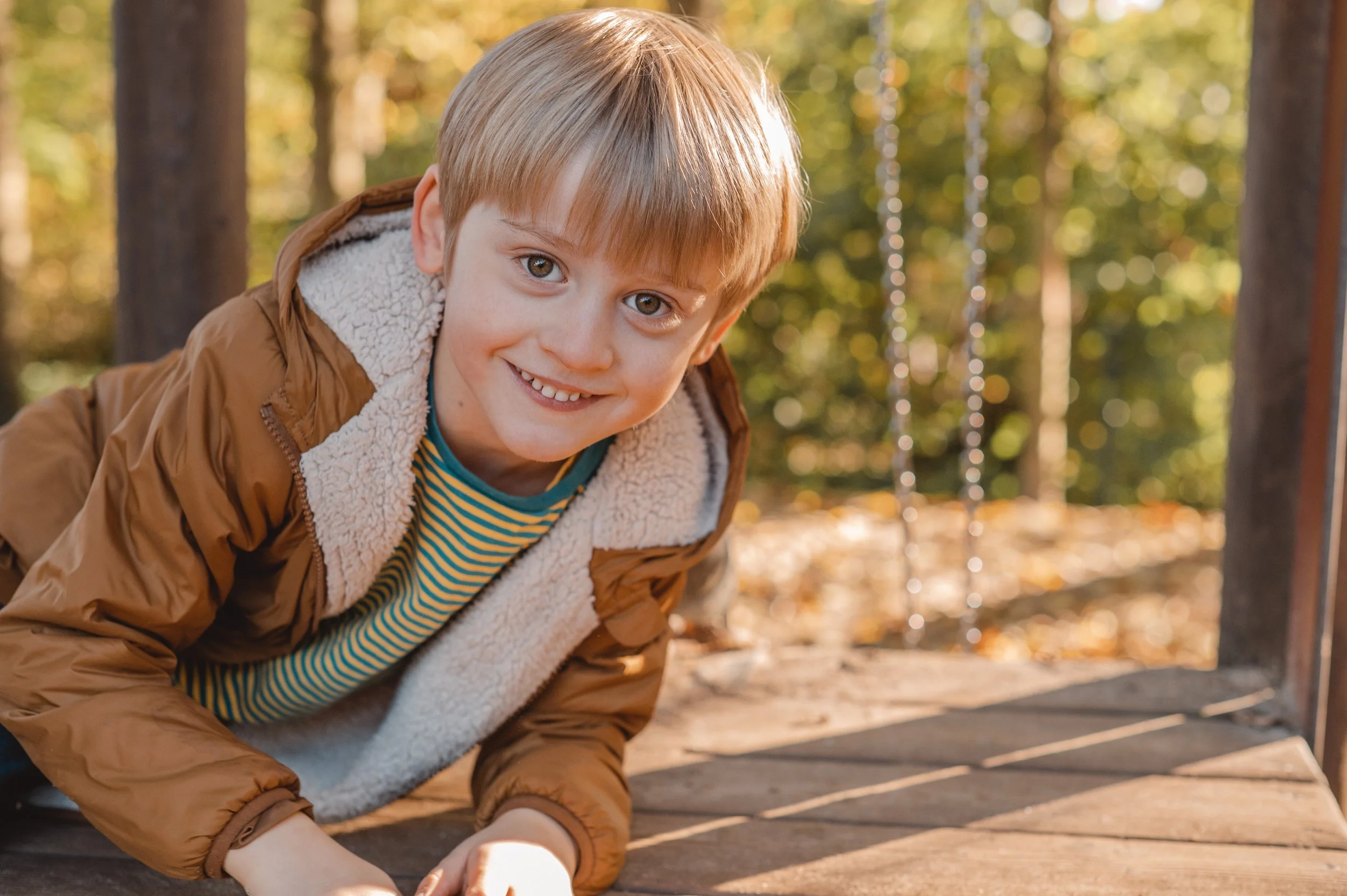 Junge stützt sich auf den Holzboden eines Klettergerüsts mit Bäumen im Hintergrund und schaut in die Kamera, auf einem Spielplatz im Herbst.