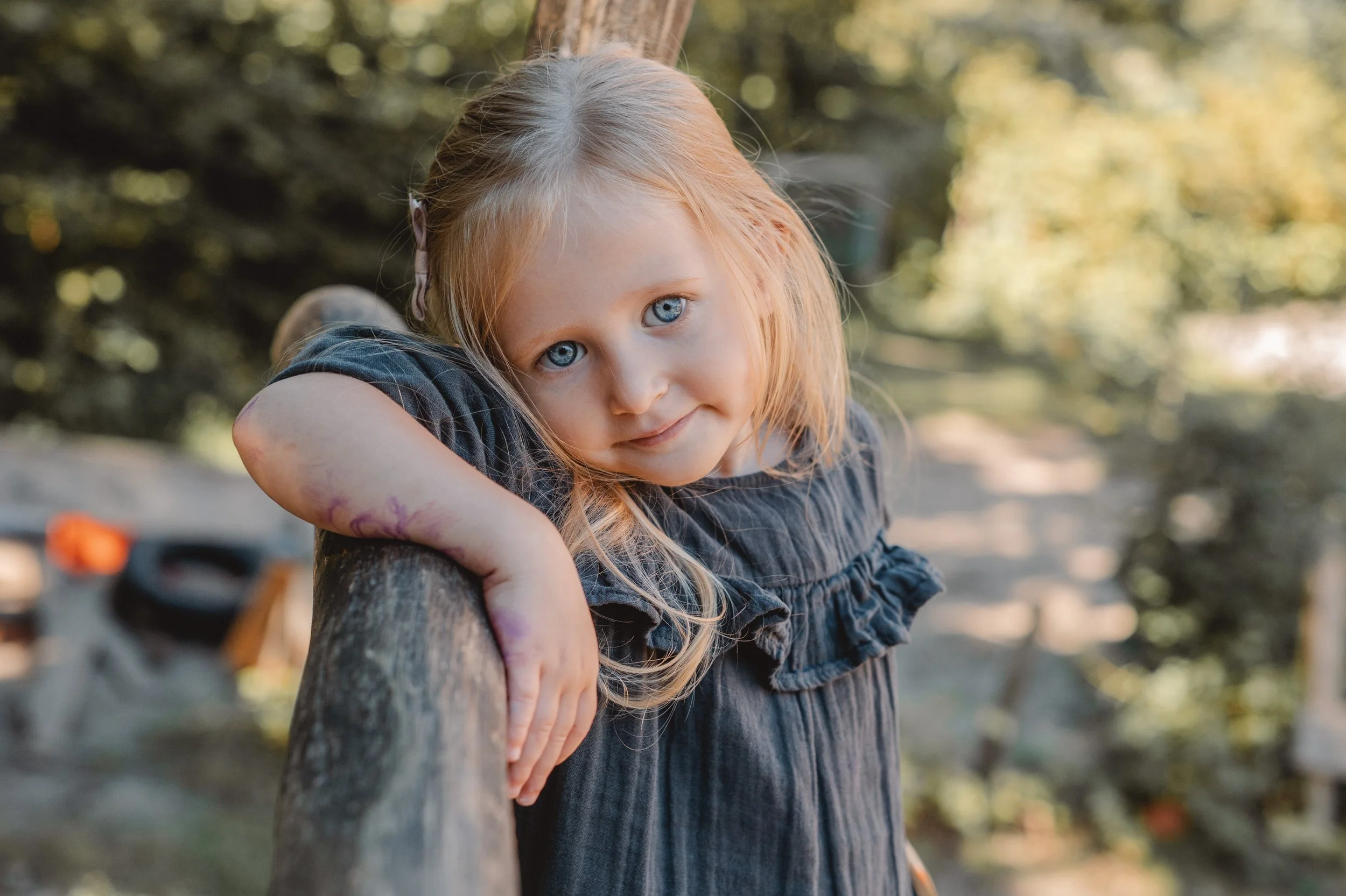 Ein junges Mädchen mit blonden Haaren und blauen Augen lehnt an einem Holzgeländer im Freien, umgeben von Bäumen.